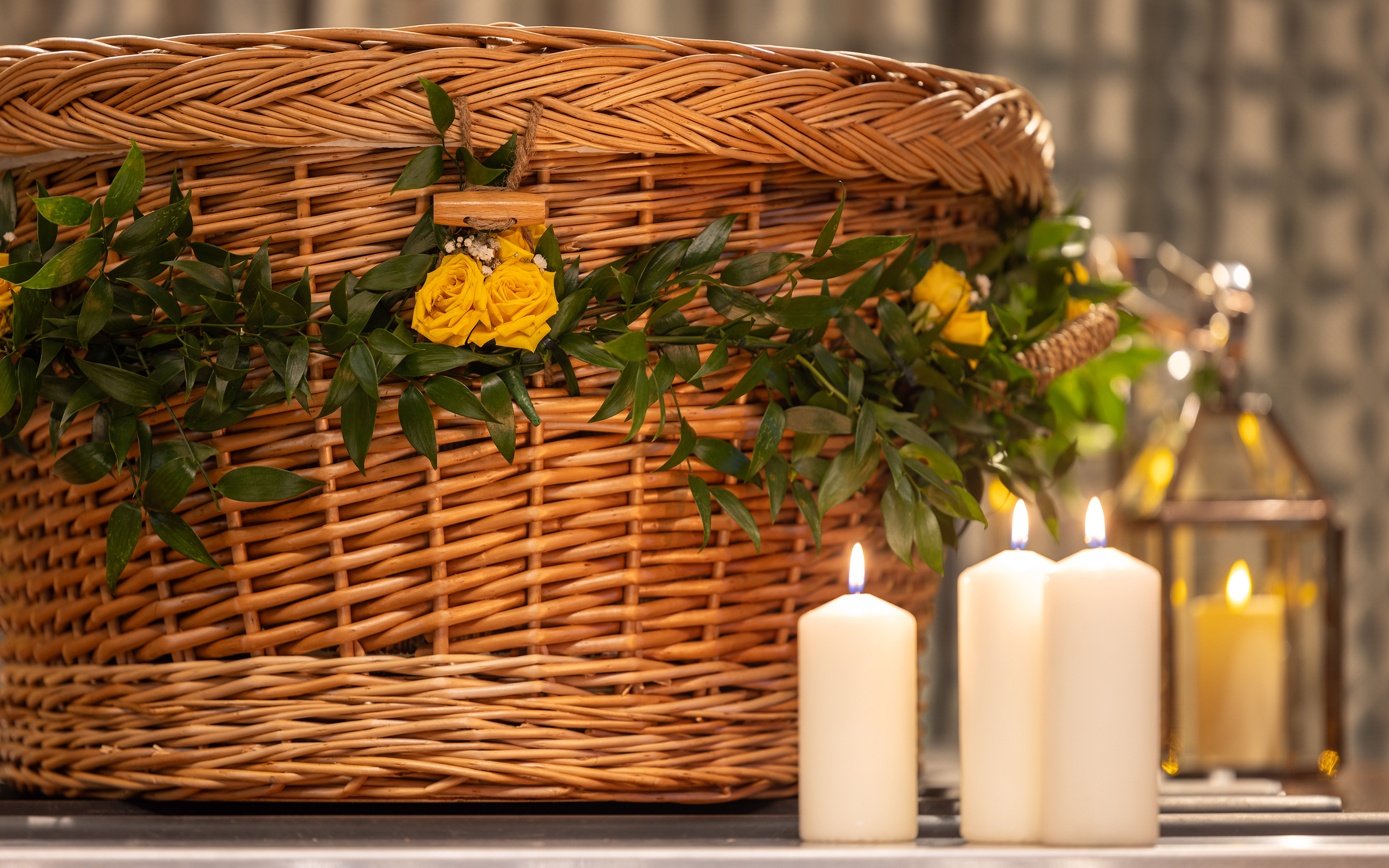 A wicker coffin dressed with foliage and yellow flowers, with lit candles and a lantern beside it.