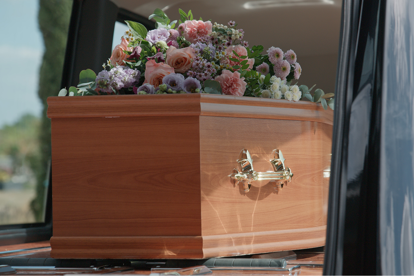 A light wooden coffin with brass handles inside a hearse, topped with a pink and purple floral spray.