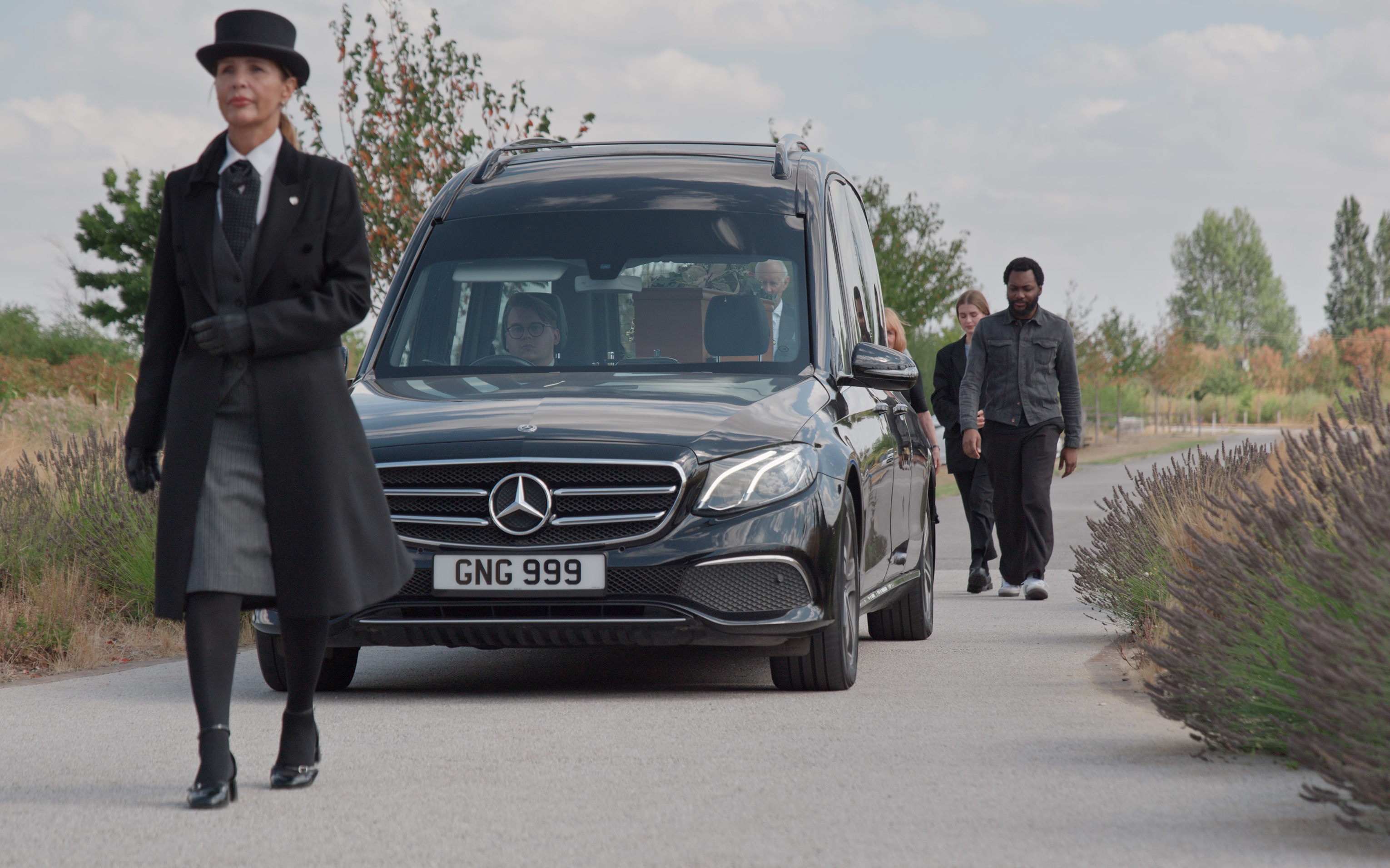A funeral director in a top hat walks ahead of a black hearse along a path.
