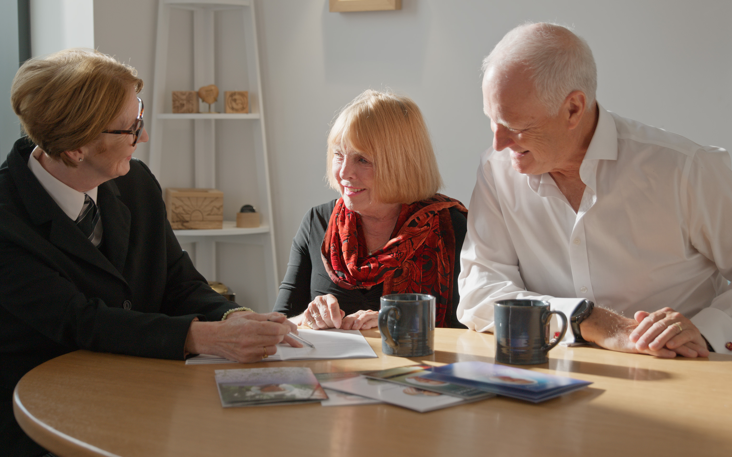 A funeral director in dark uniform talks with a woman and an older man across a table.