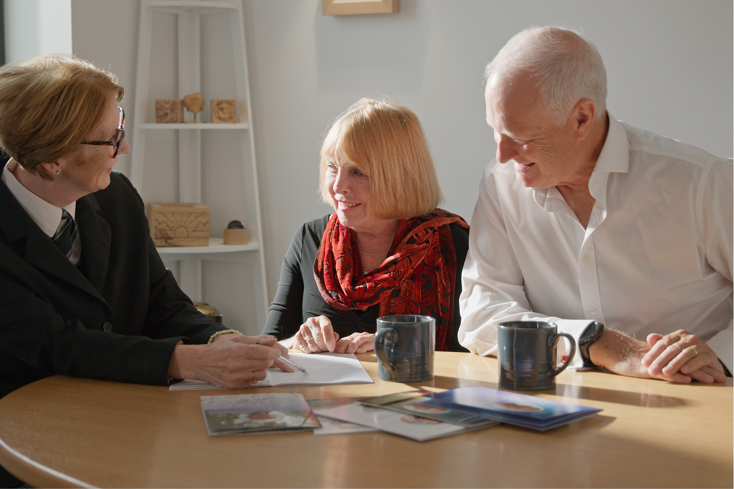An older couple smile as a funeral arranger writes notes at a table with brochures.