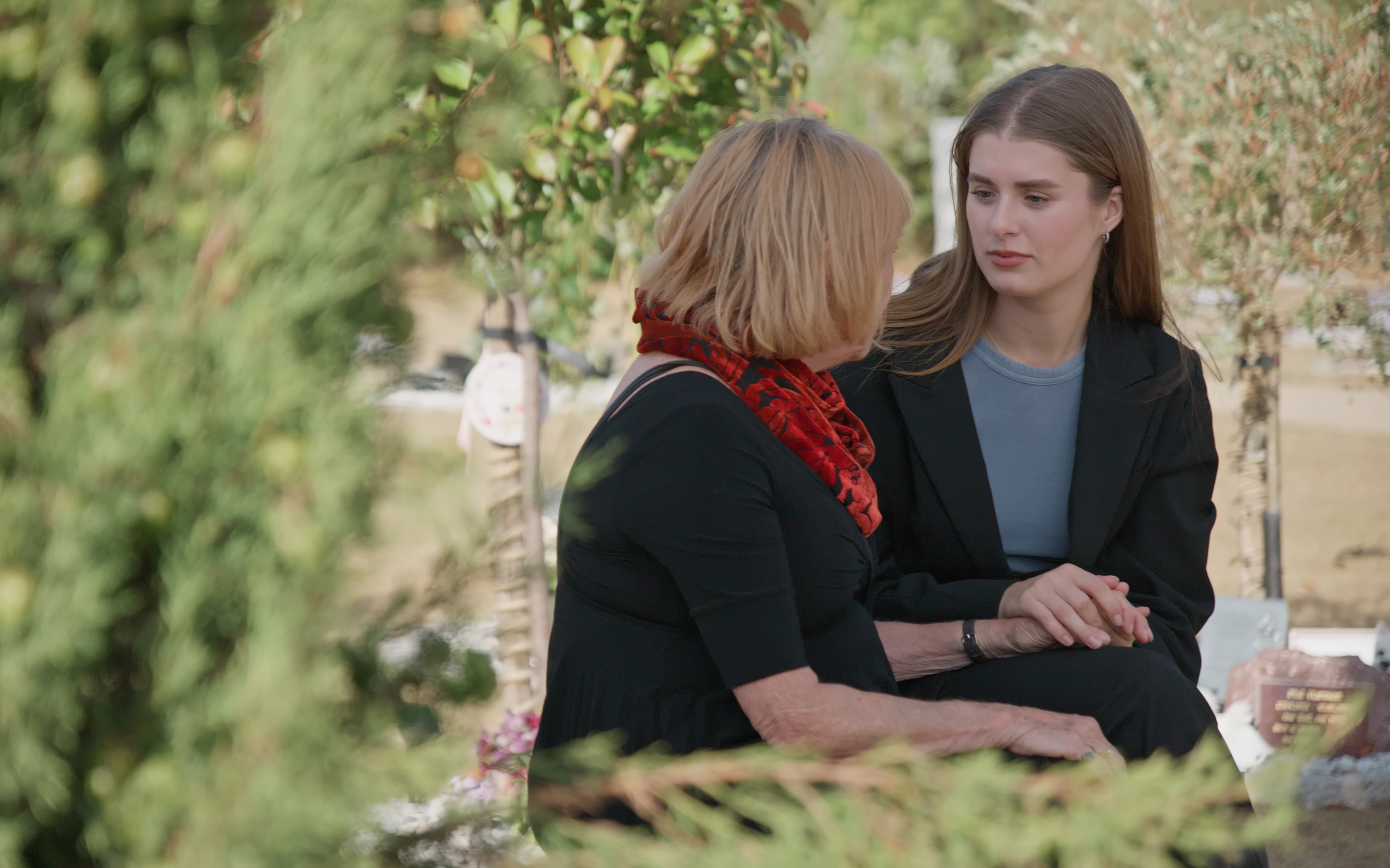 An older woman and a younger woman sit talking together in a sunlit garden.
