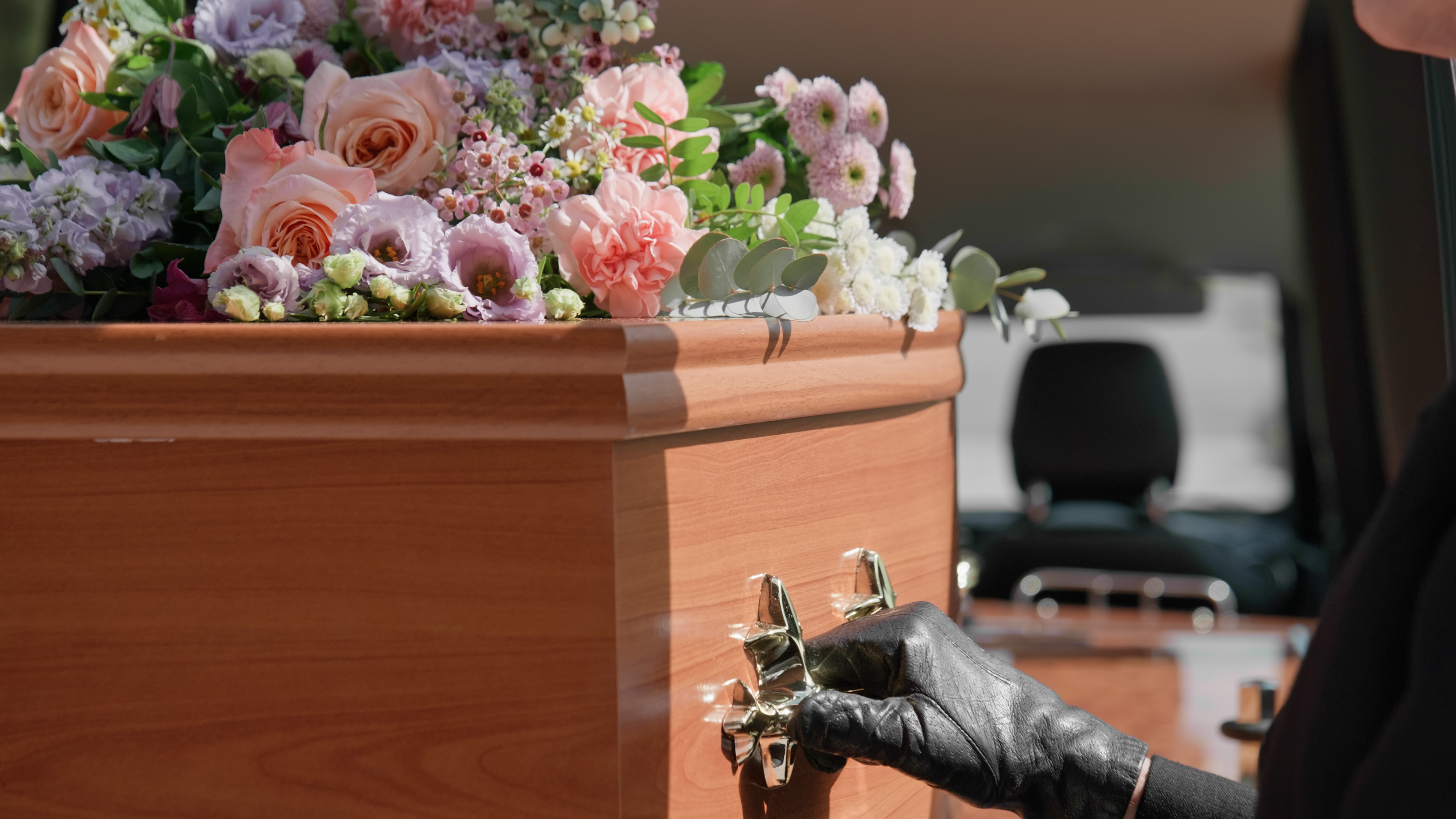 A gloved hand rests on the brass handle of a flower-topped wooden coffin in a hearse.