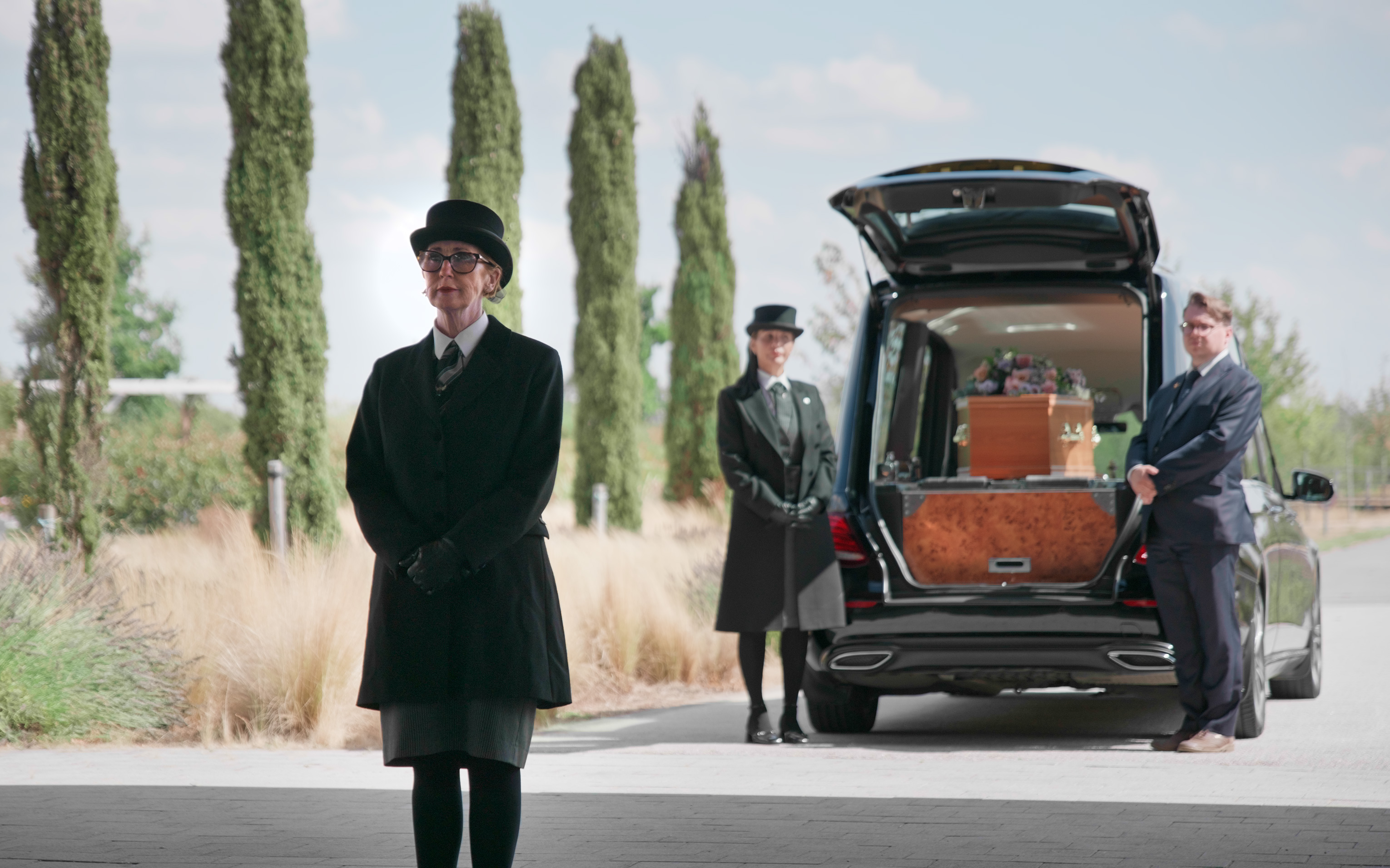 Funeral directors in top hats standing beside an open hearse holding a wicker coffin.