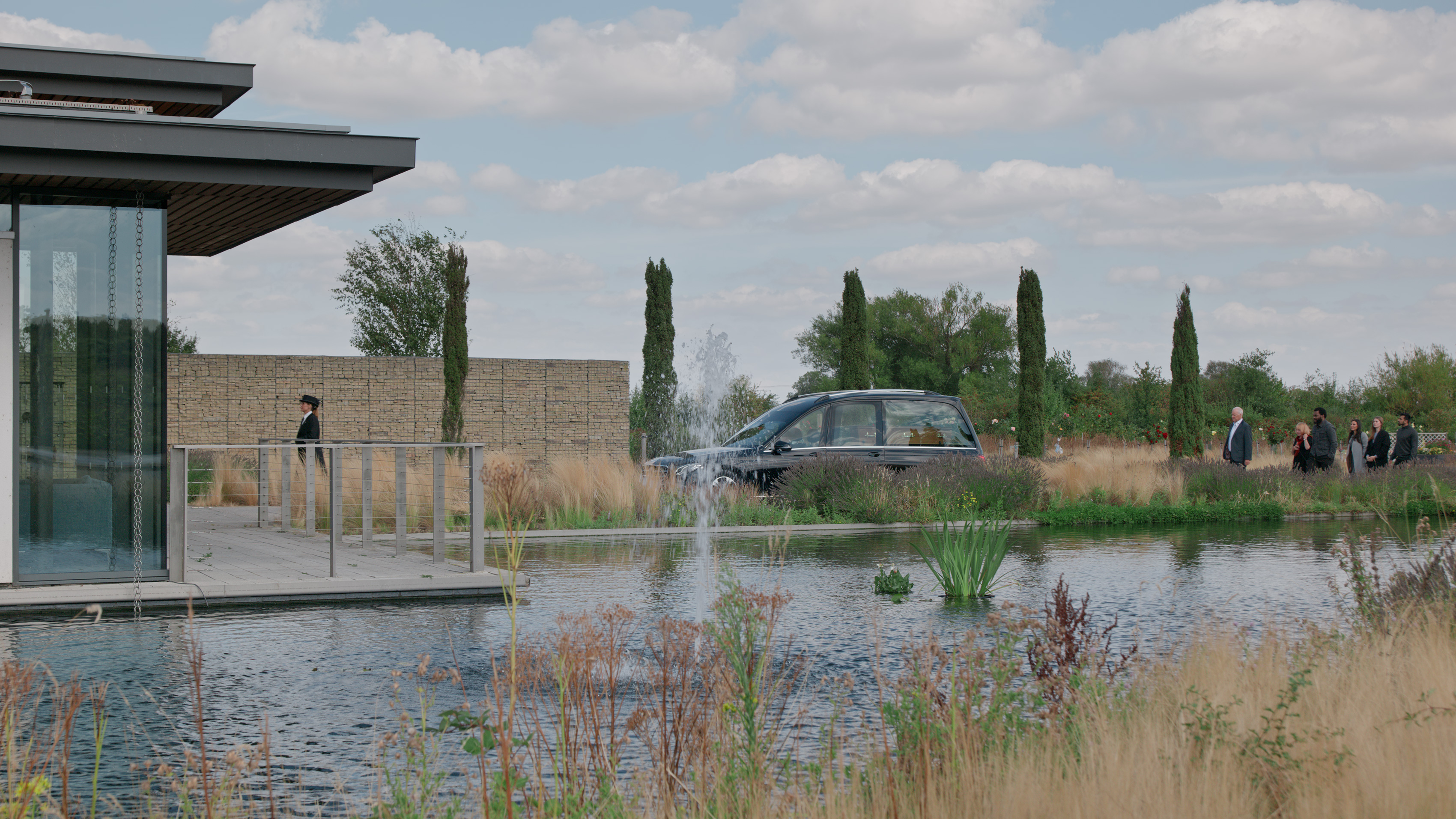 A glass-fronted modern building beside a landscaped lake, with a hearse and mourners outside.