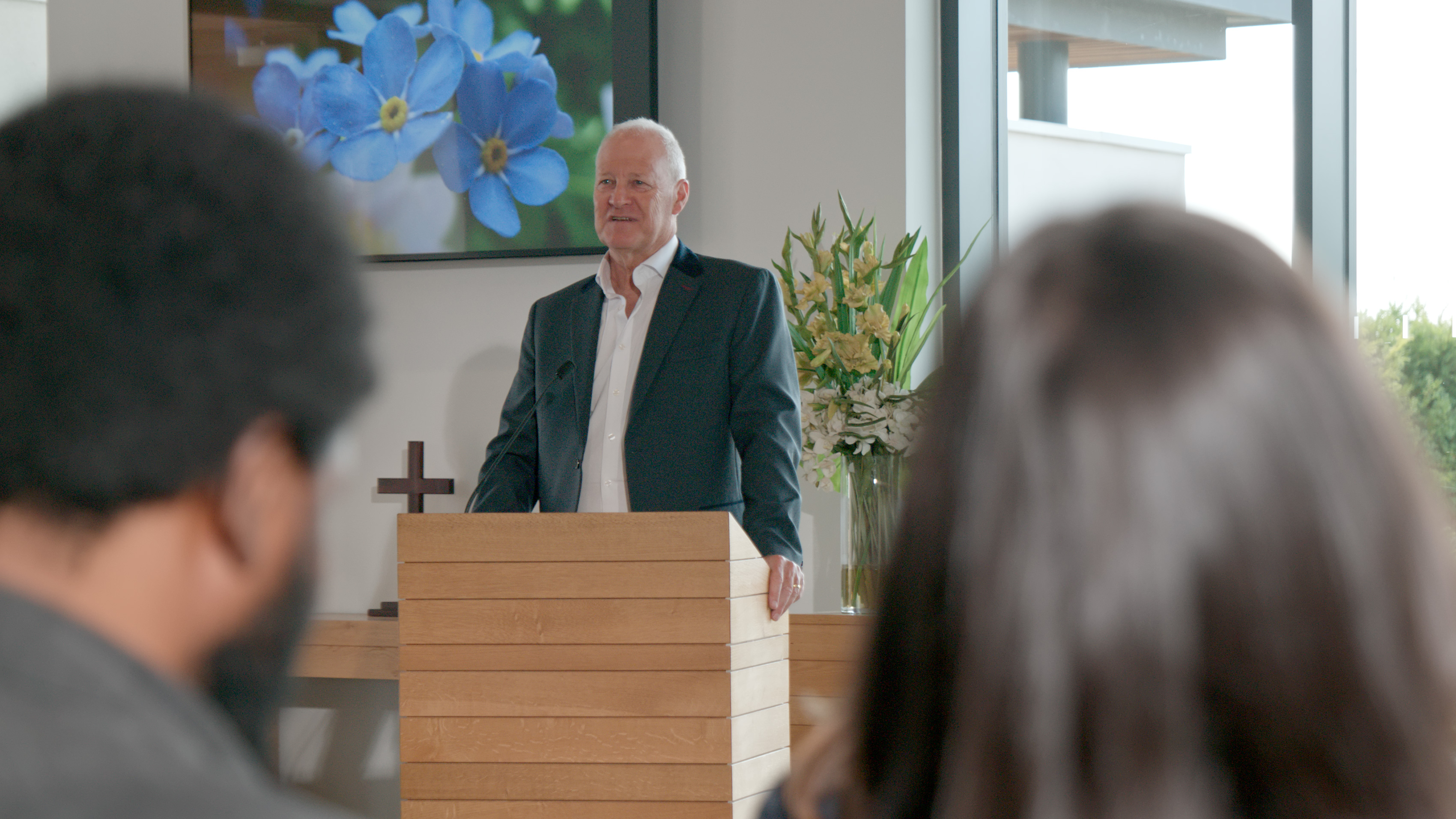 A man stands at a wooden lectern addressing a service, seen over the shoulders of two mourners.