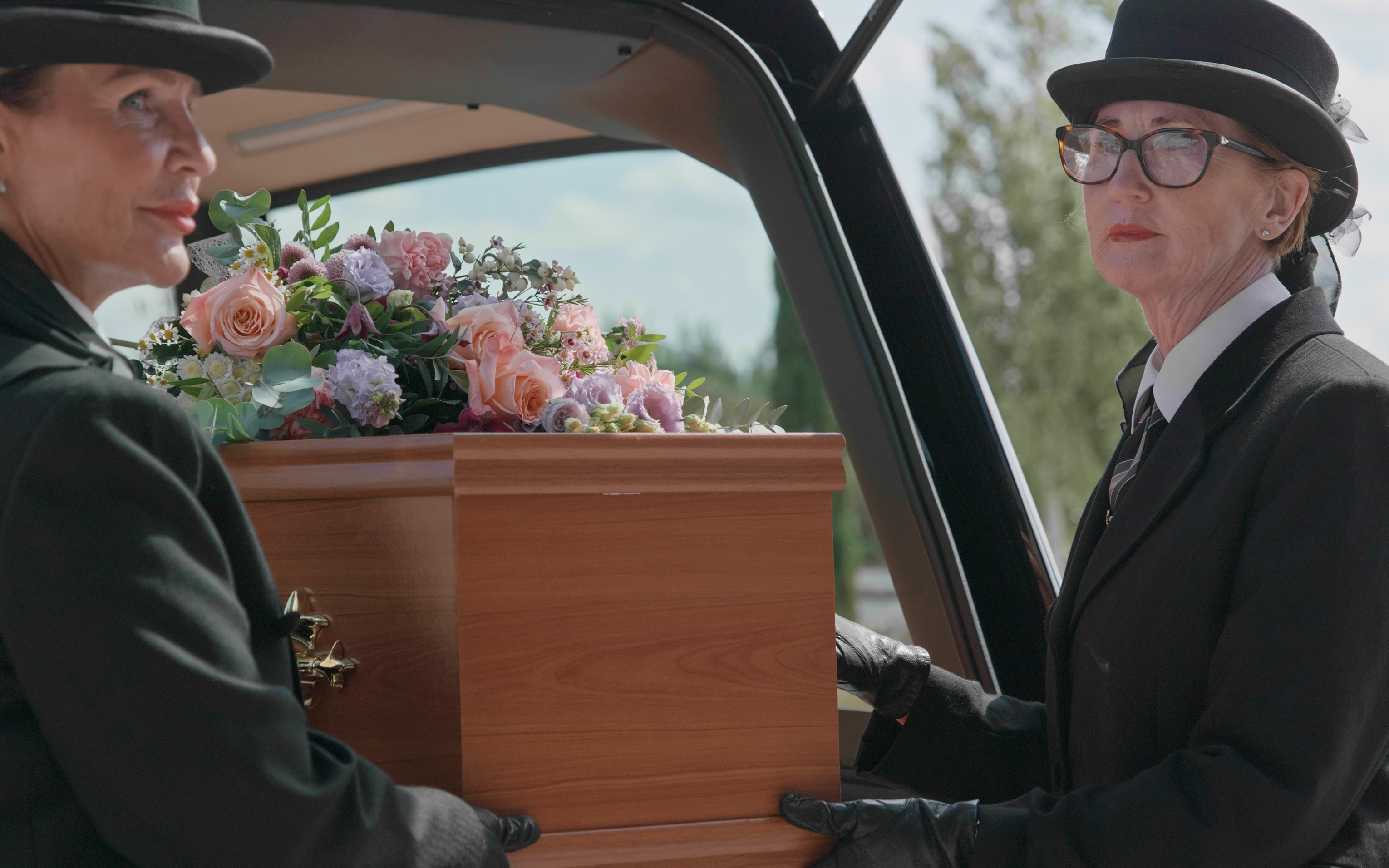 Two Dignity funeral directors in top hats at the open back of a hearse, with a coffin dressed in pink and lilac flowers.