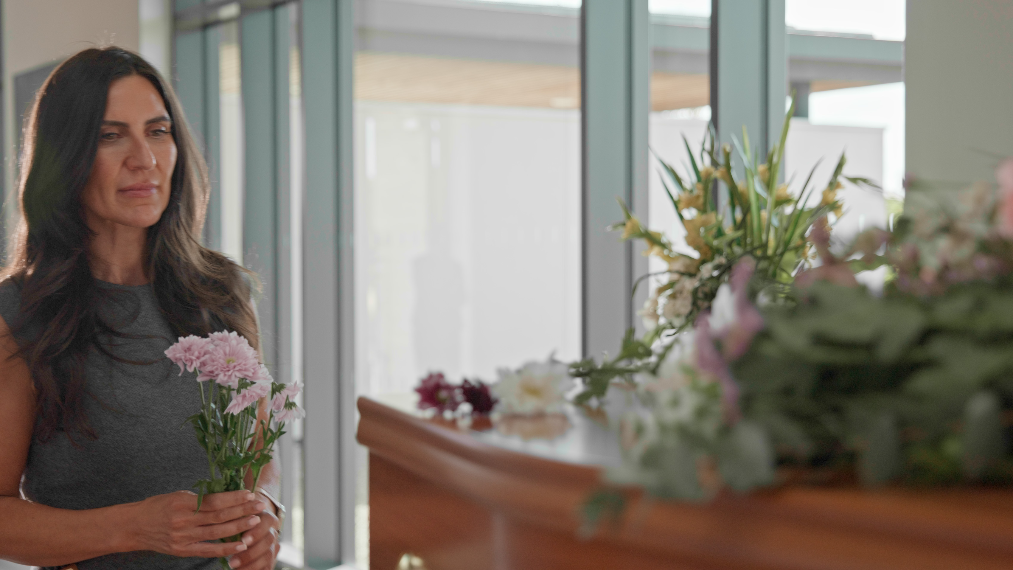 A woman holding pink carnations beside a wooden coffin dressed with flowers in a bright room.
