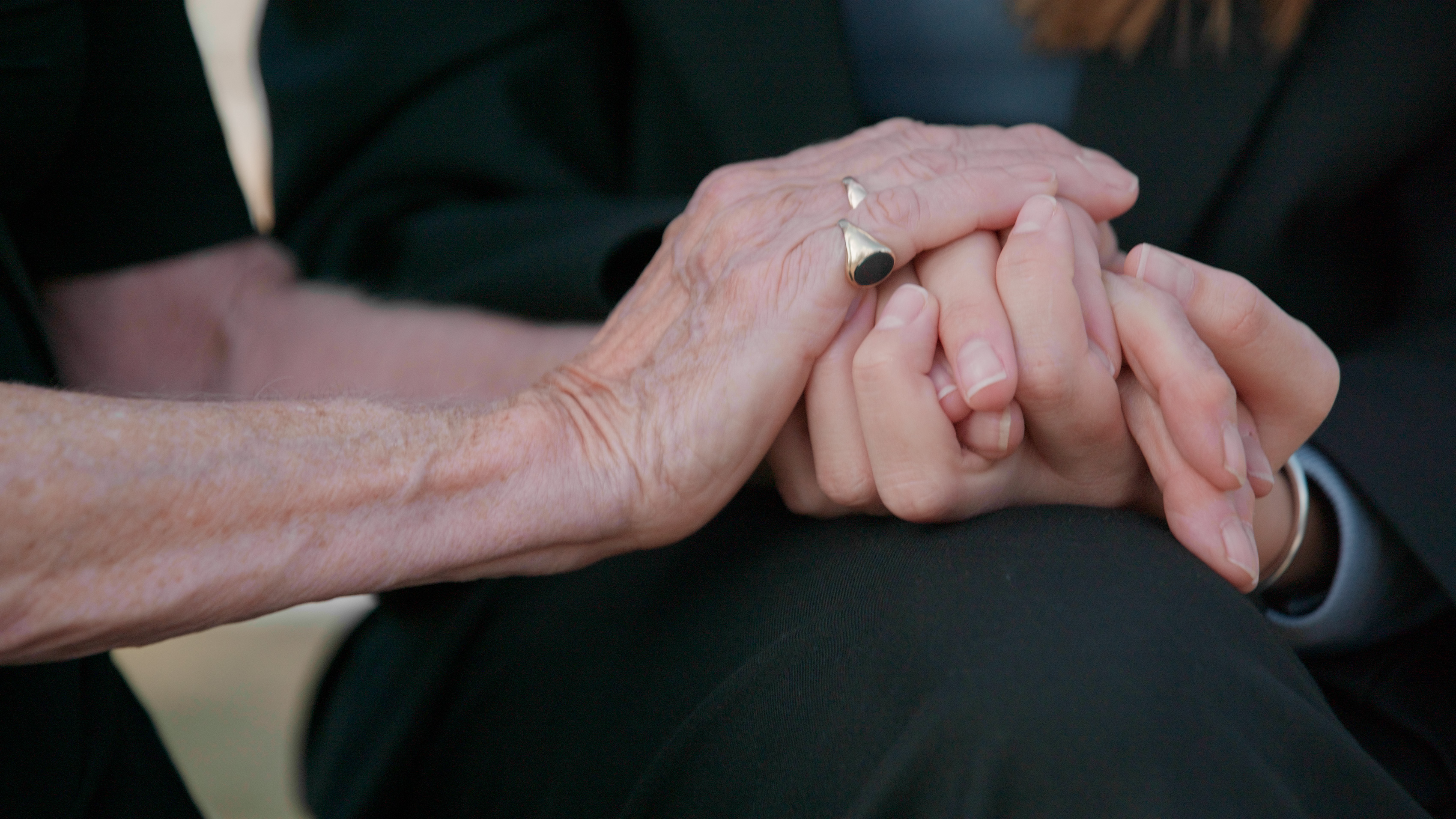 Close-up of an older person's hands clasped in their lap, wearing a wedding ring.