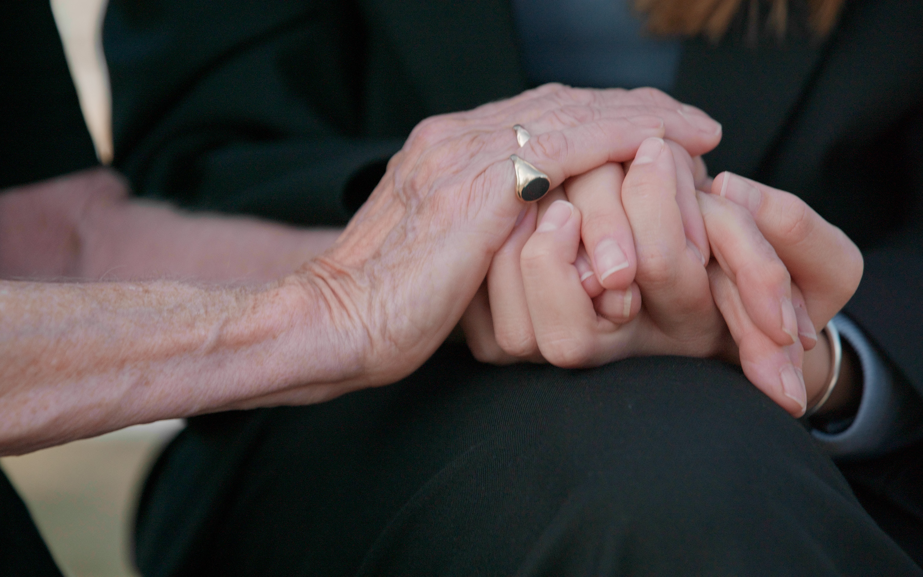 Close-up of two people's hands held together on a lap, one older hand wearing a ring.
