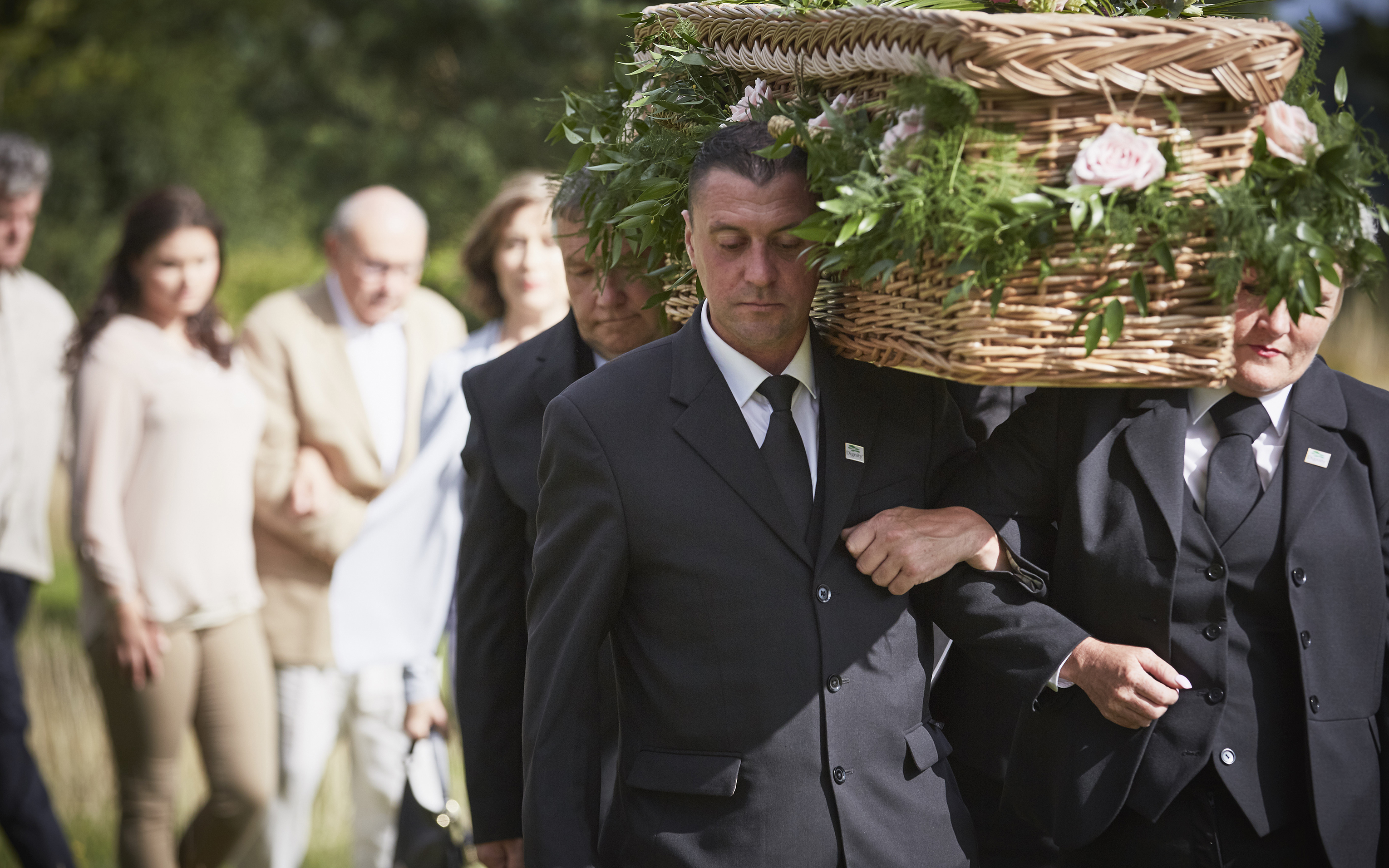 Pallbearers in dark suits carrying a wicker coffin covered with foliage, with mourners following behind.