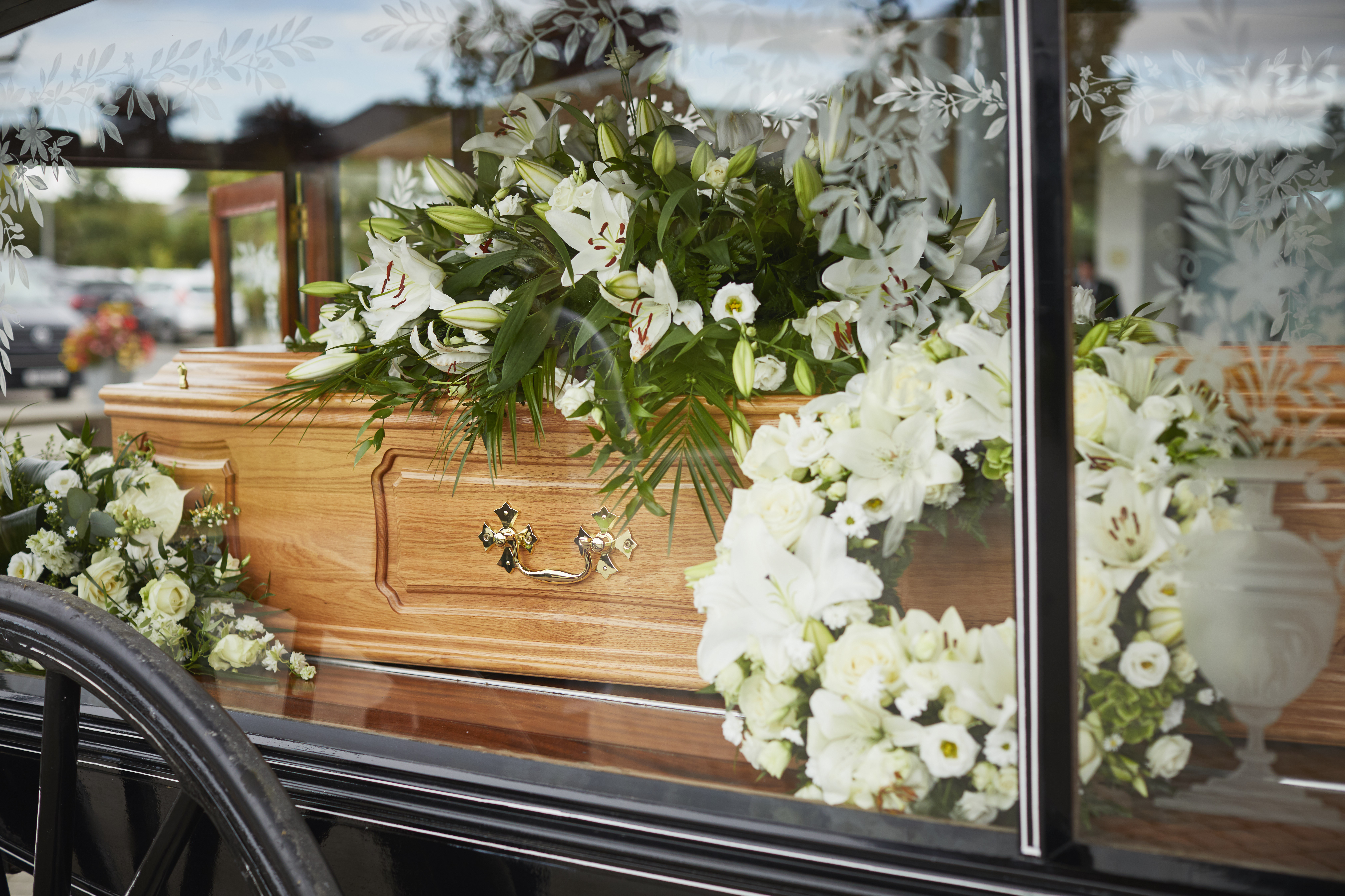 A wooden coffin covered with white lilies, seen through the etched glass side of a hearse.