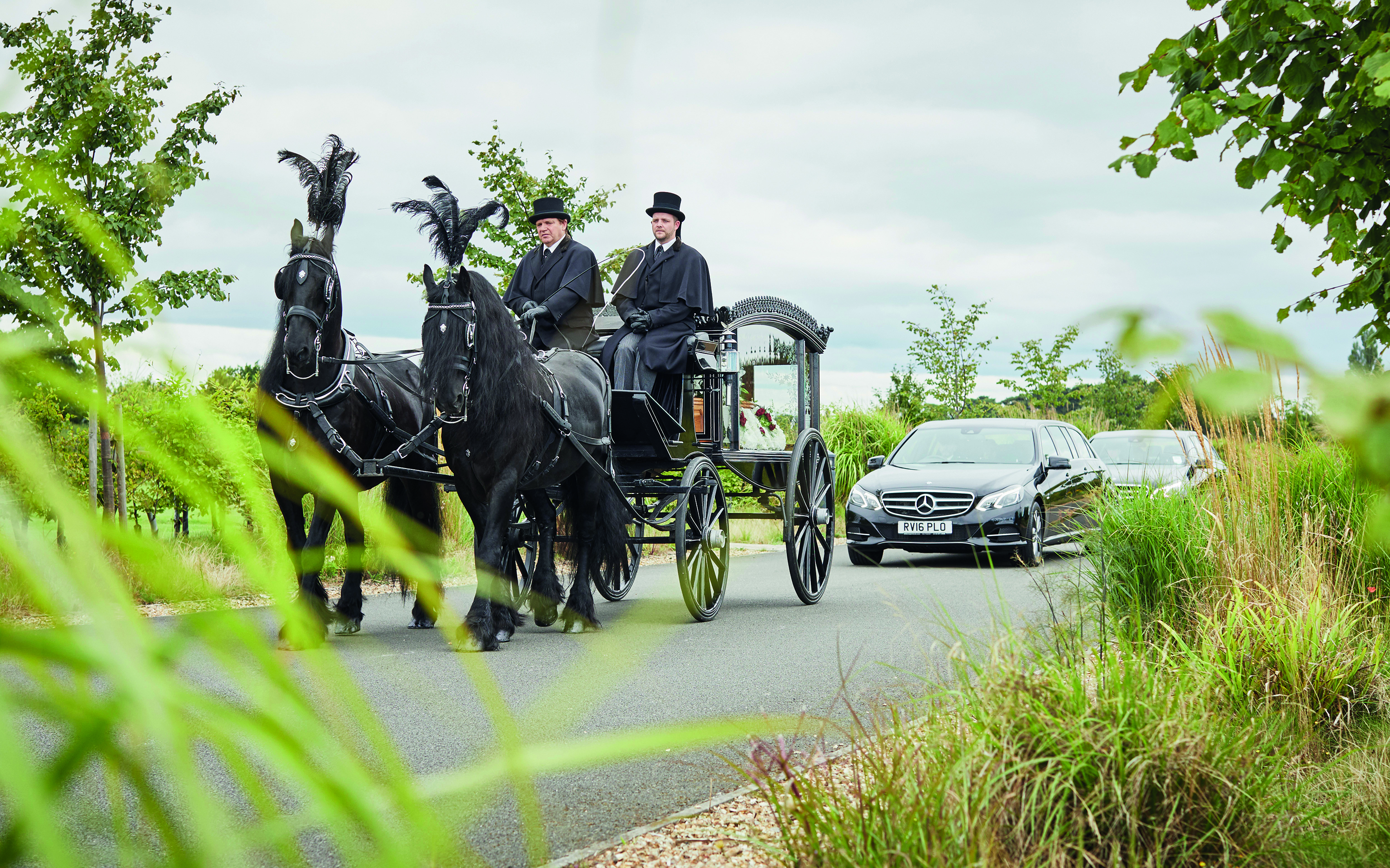 A horse-drawn hearse with two black plumed horses and a top-hatted driver, followed by a car.