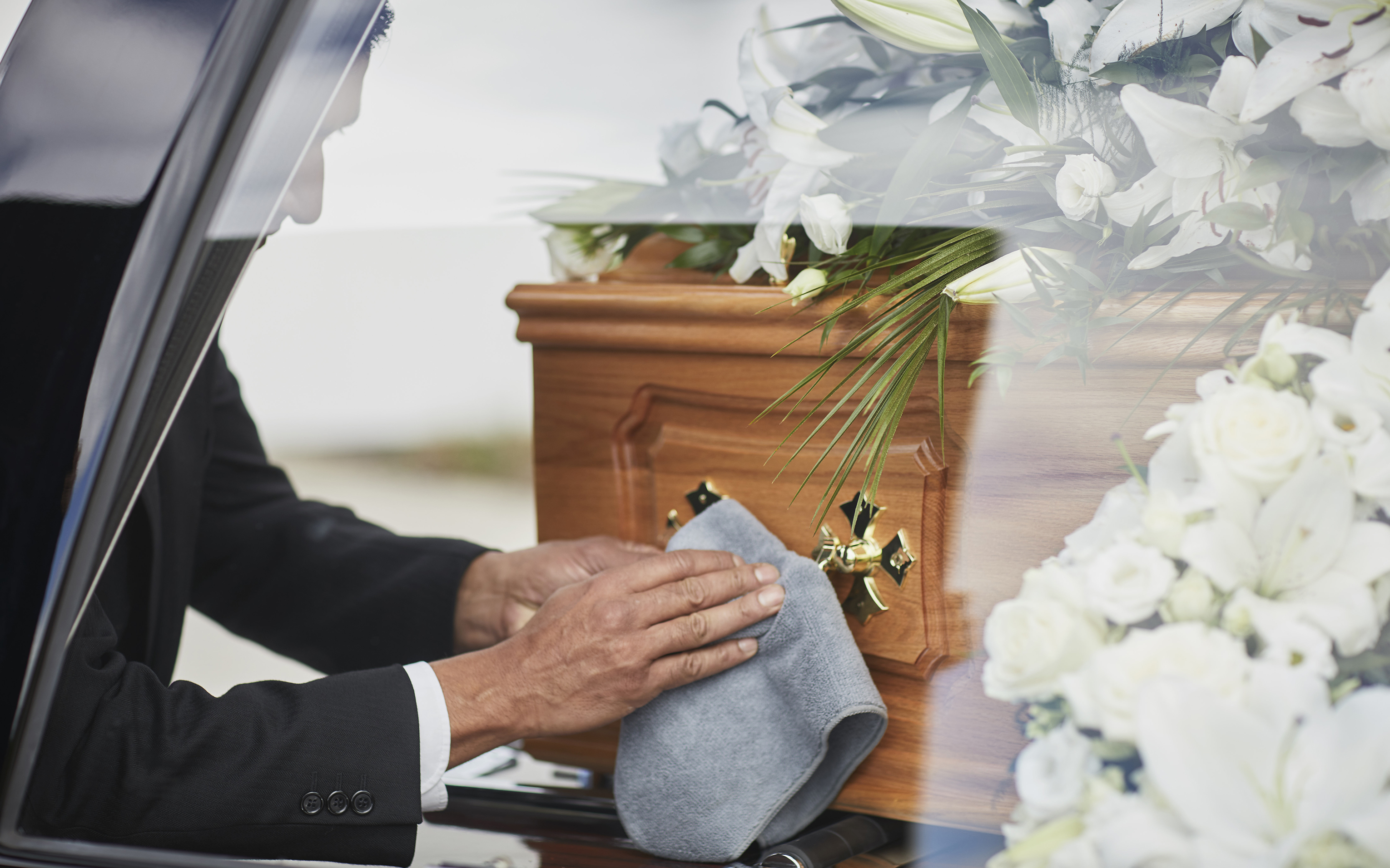 A man in a dark suit polishes a wooden coffin with a cloth, white lilies alongside.