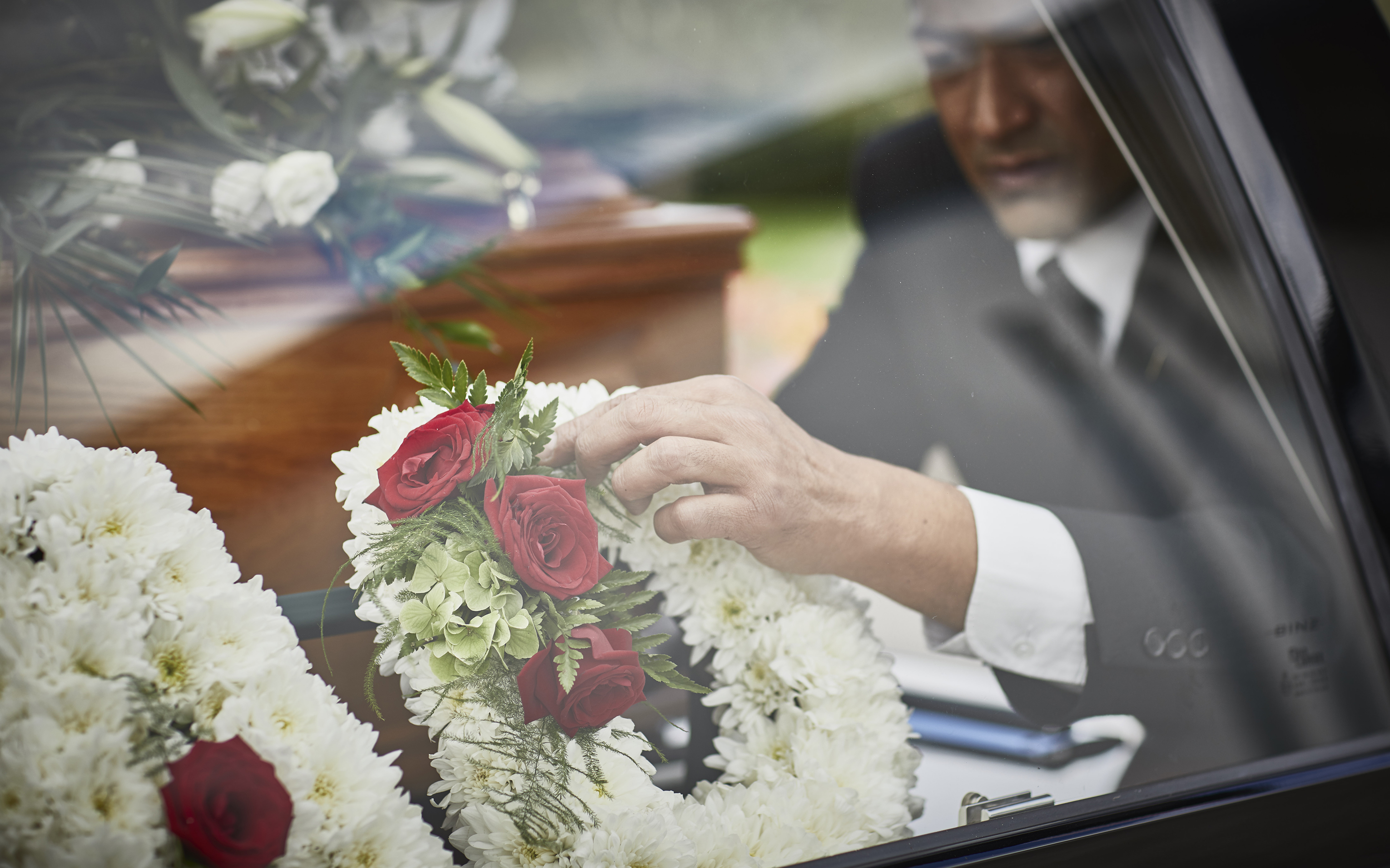 A funeral director arranging a wreath of white chrysanthemums and red roses beside a hearse.