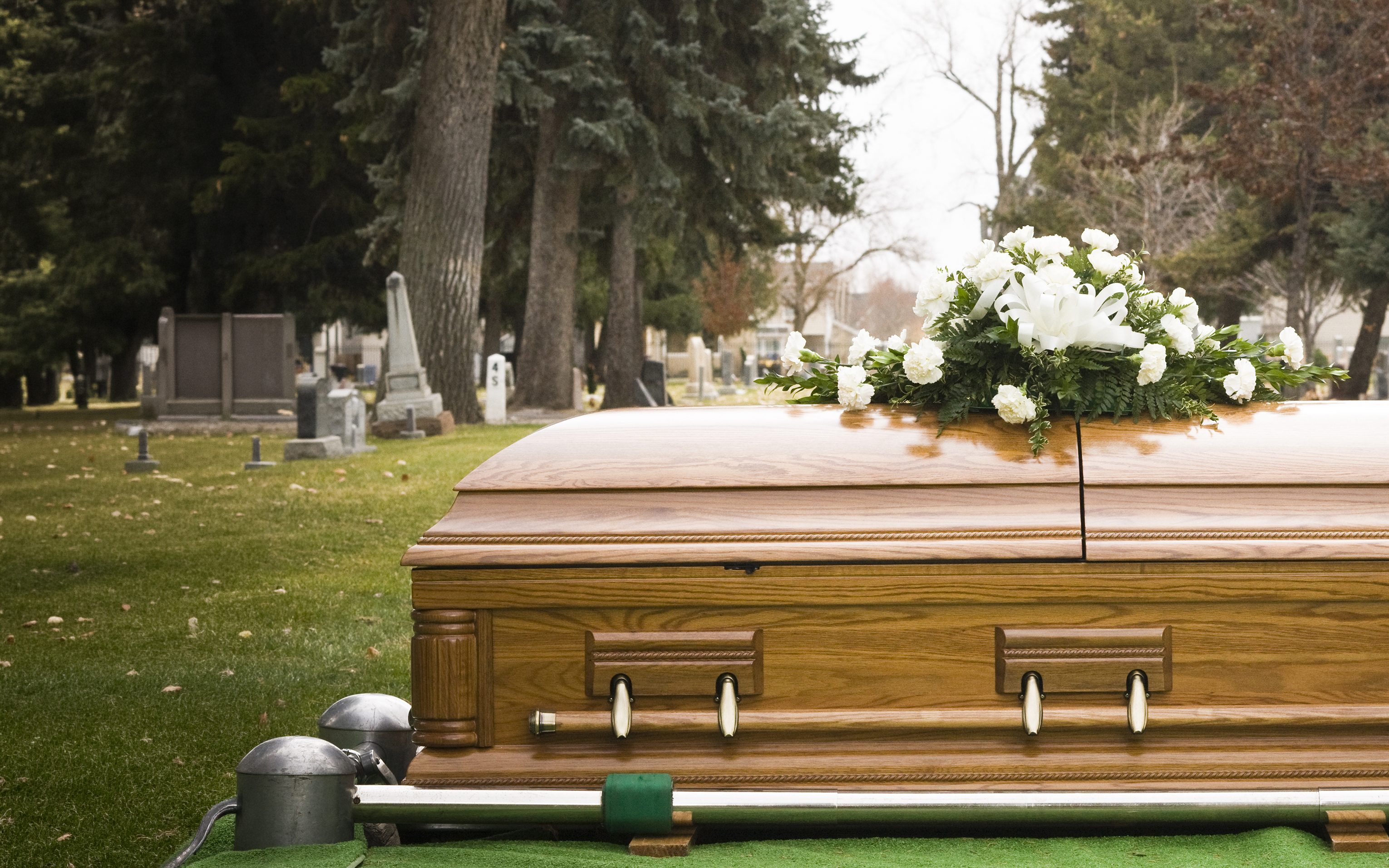 A wooden coffin with white flowers on top, beside a grave in a cemetery.