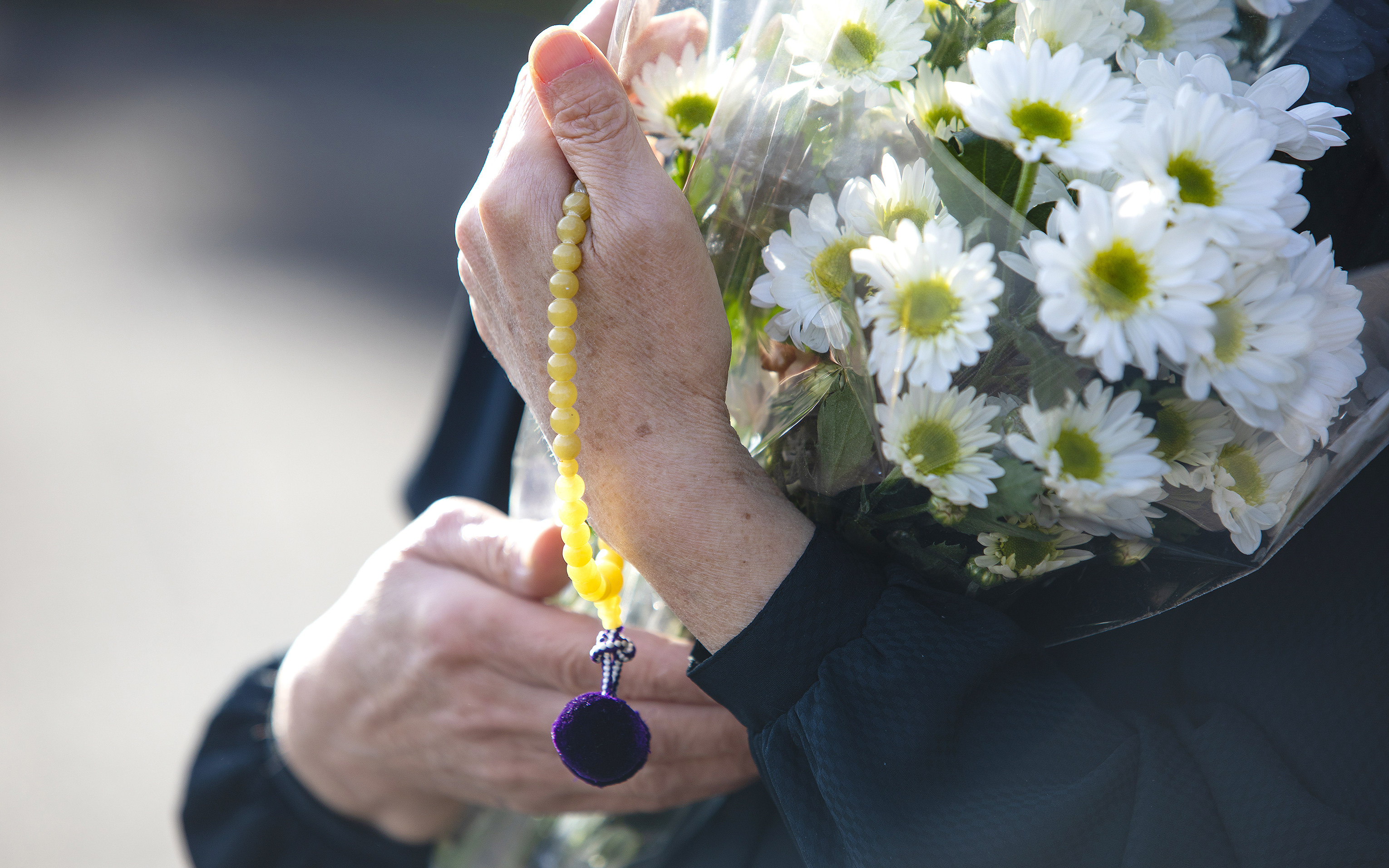 Hands holding a wrapped bunch of white chrysanthemums and a string of yellow prayer beads.