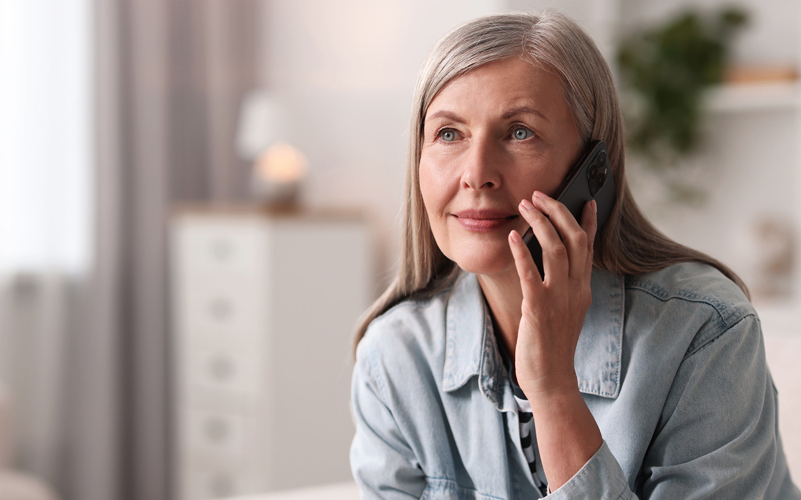 An older woman with grey hair holds a mobile phone to her ear at home.