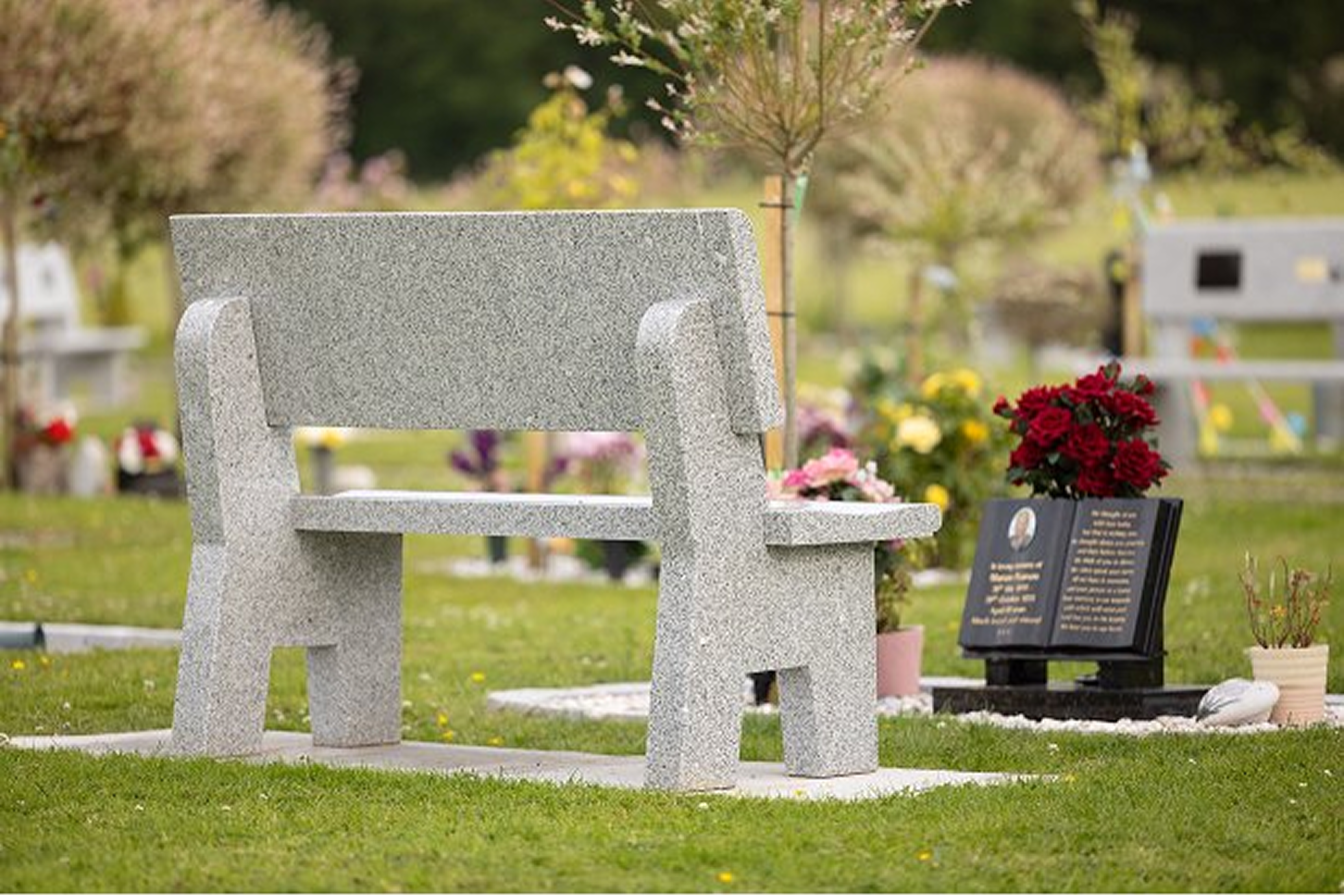 A grey granite memorial bench on a lawn, beside a black memorial plaque and red flowers.