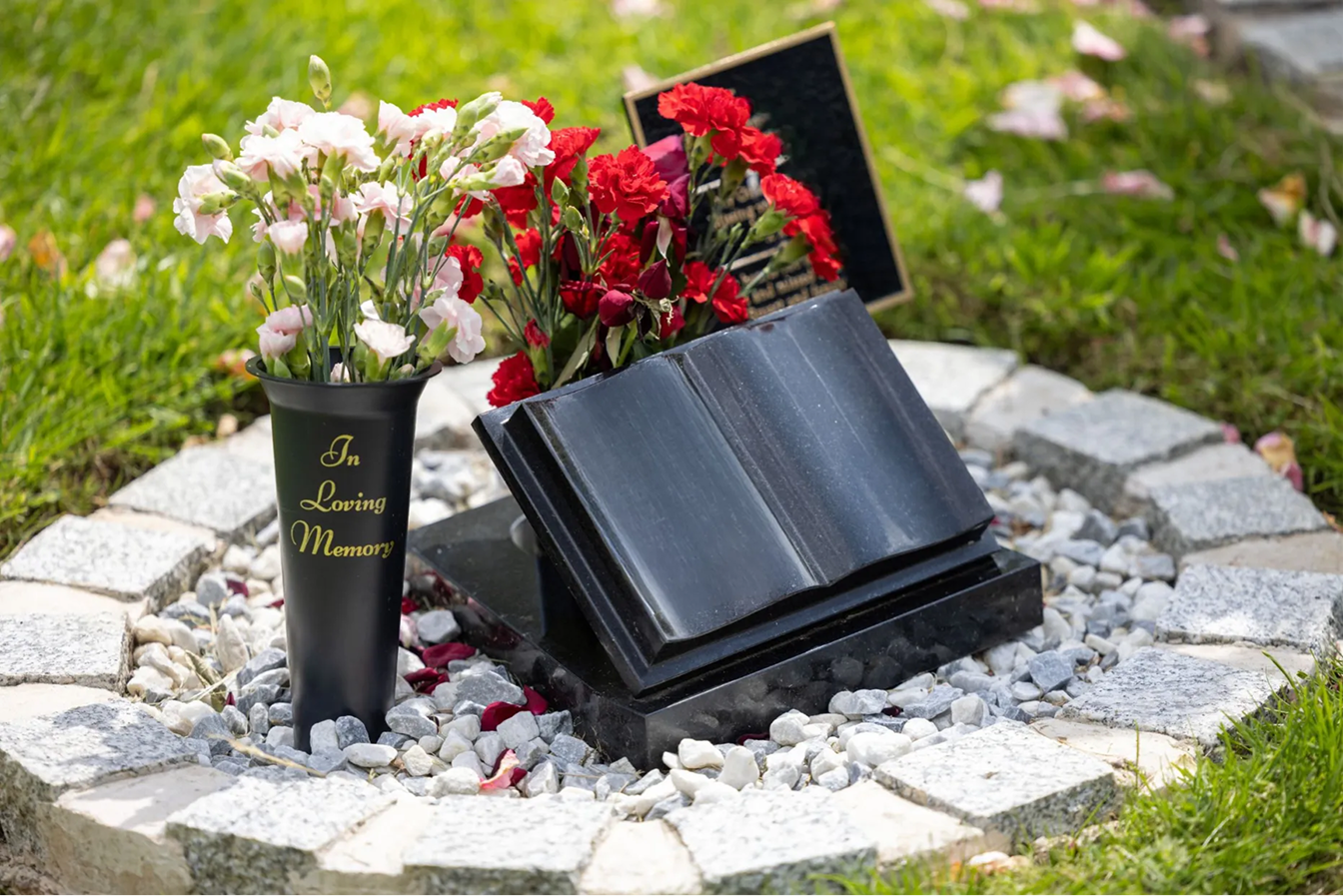 A black granite open-book memorial on a stone-edged grave with red and white carnations.