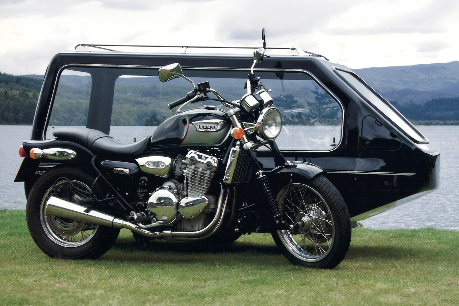 A black motorcycle hearse with a glass-sided carriage, parked on grass beside a lake.