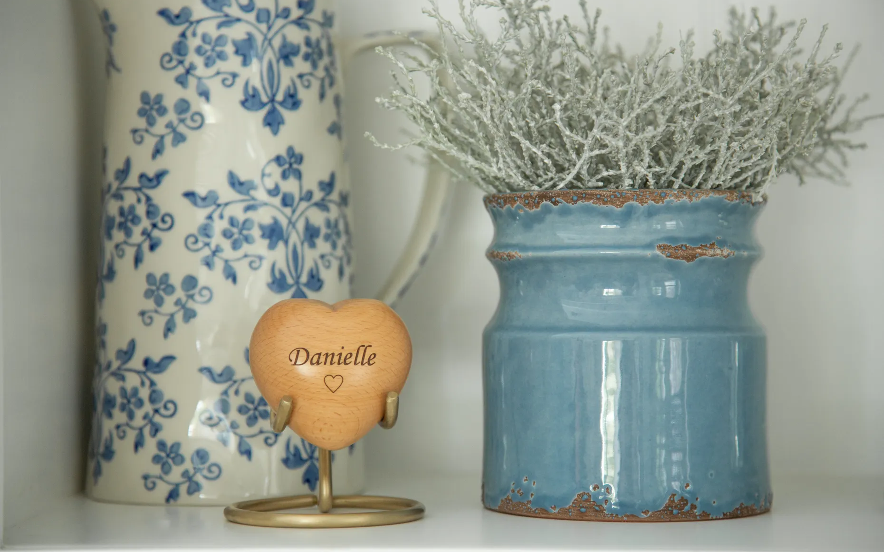 A wooden heart keepsake engraved Danielle on a brass stand, beside a patterned jug and blue pot.
