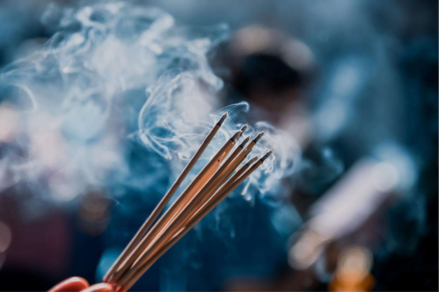 A hand holding a bundle of lit incense sticks, smoke rising against a dark background.