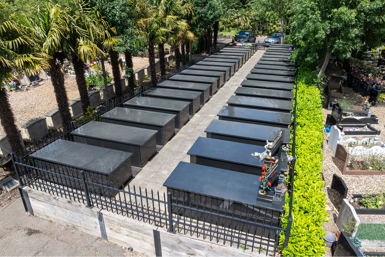 Rows of polished black granite burial vaults behind iron railings, with trees and hedging around them.