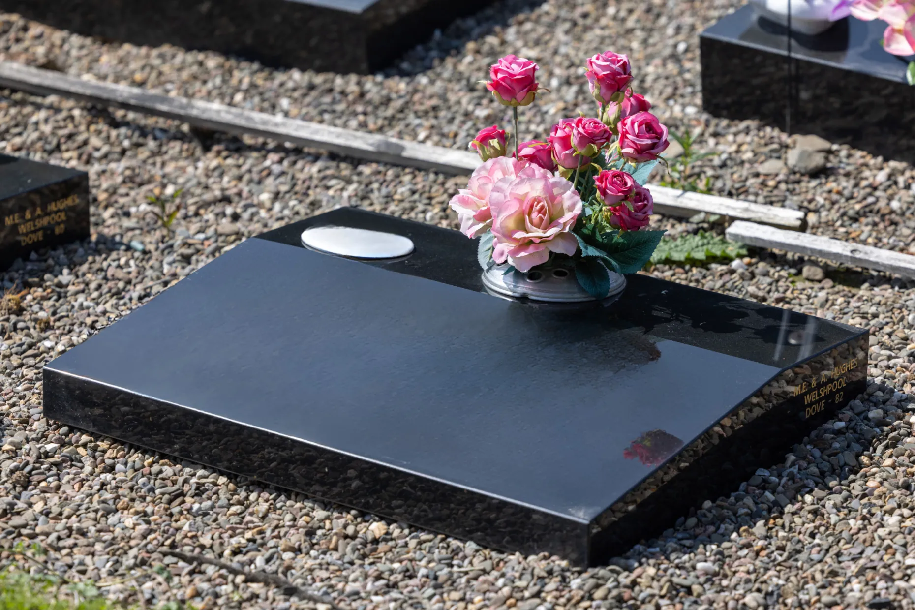 A black granite memorial with pink roses laid on the gravel beside it.