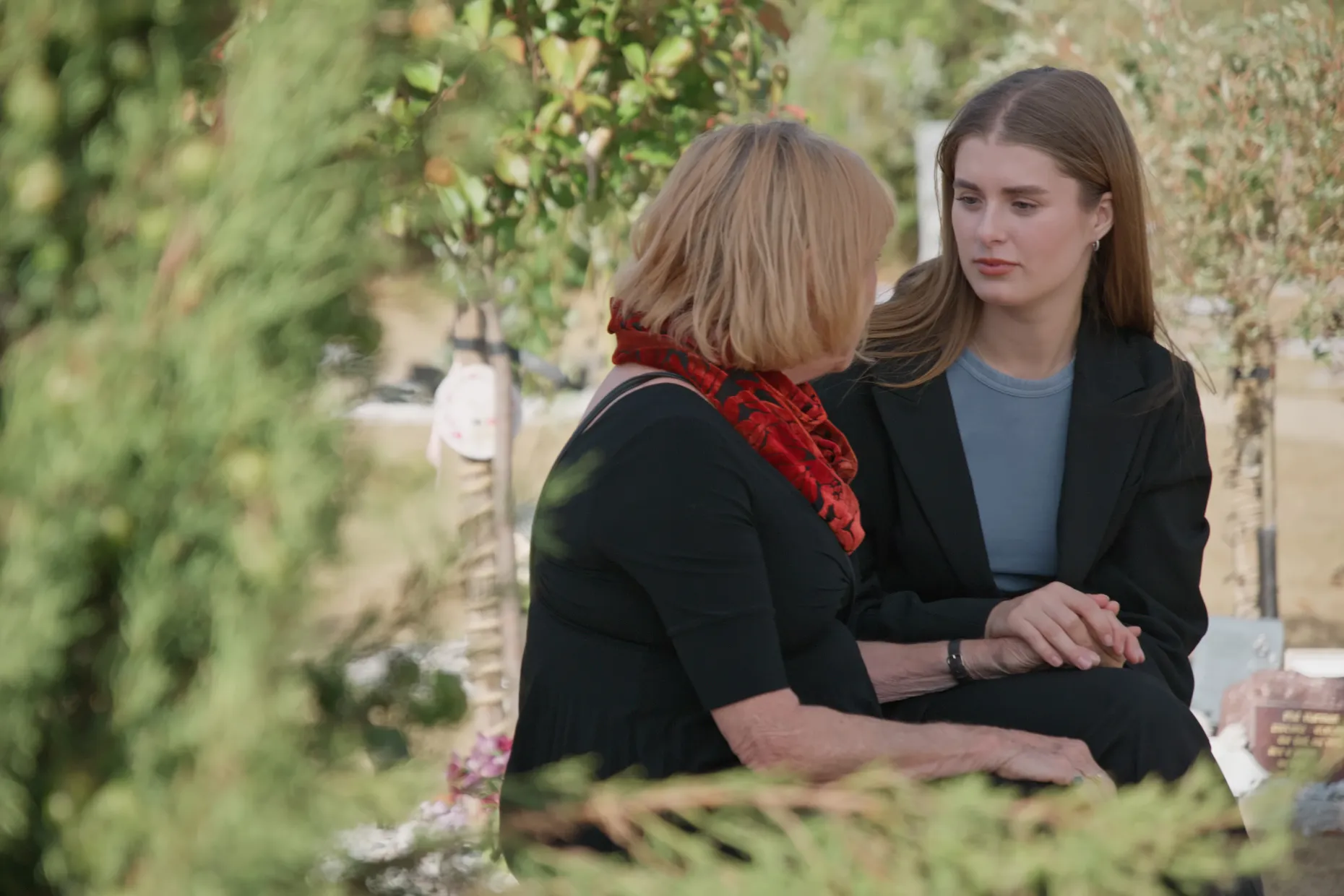 An older woman and a younger woman sit talking together in a sunlit garden.