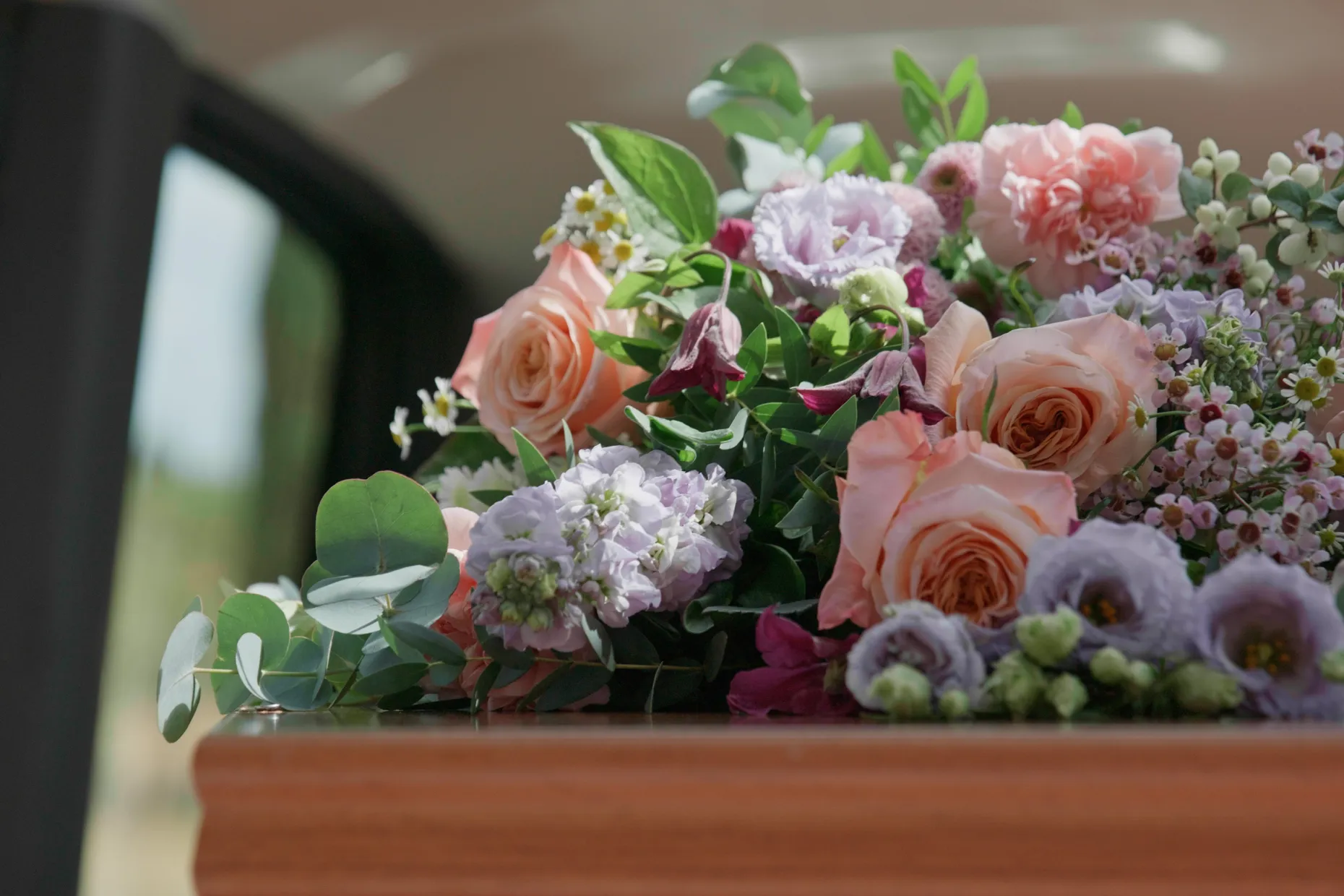 A wooden coffin in the back of a hearse, with peach roses and purple flowers on top.