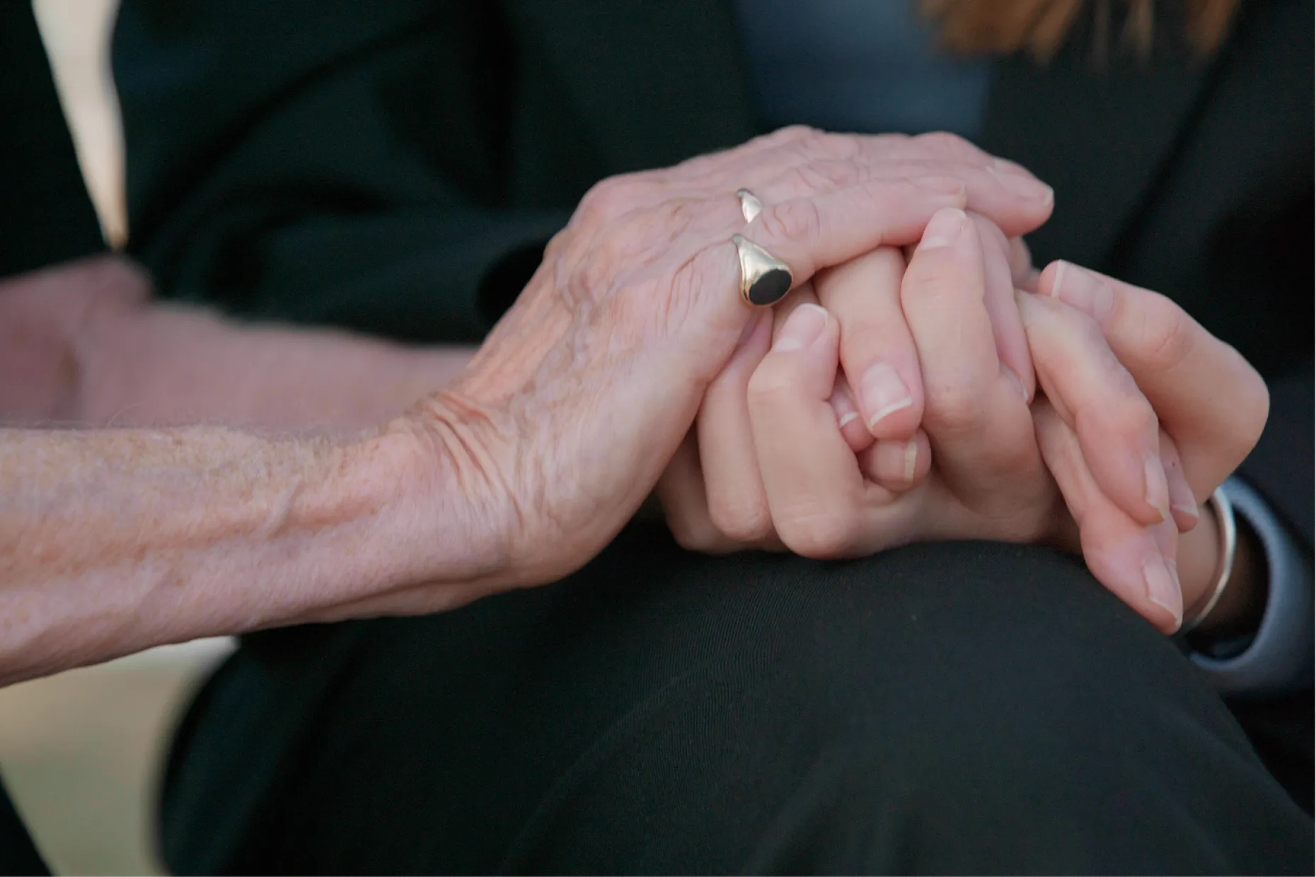 Close up of an older person's clasped hands wearing rings, dressed in dark clothing.