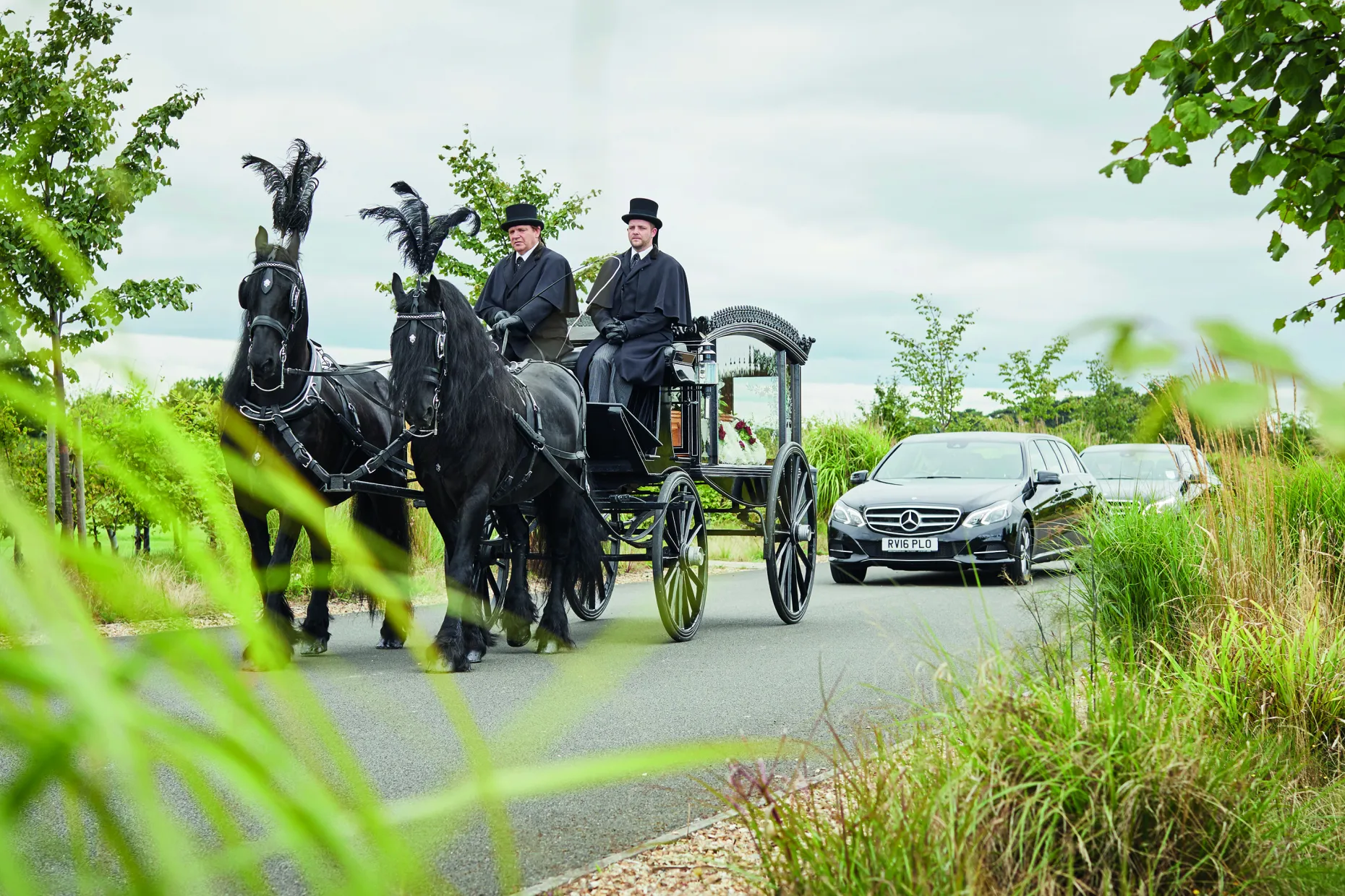 A horse-drawn hearse with two black plumed horses and a top-hatted driver, followed by a car.