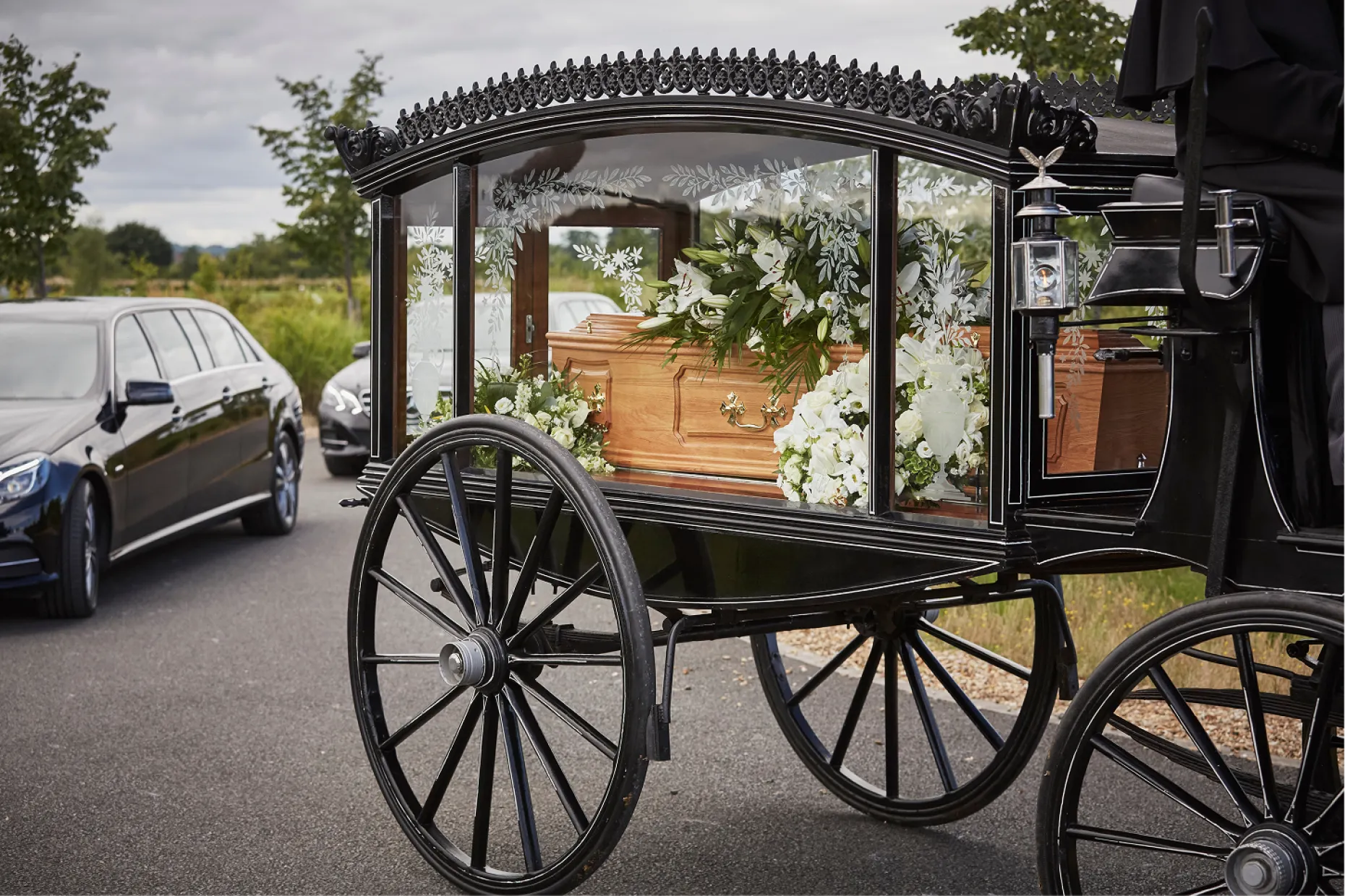 A glass sided funeral carriage carrying a coffin and white flowers, parked on a road.