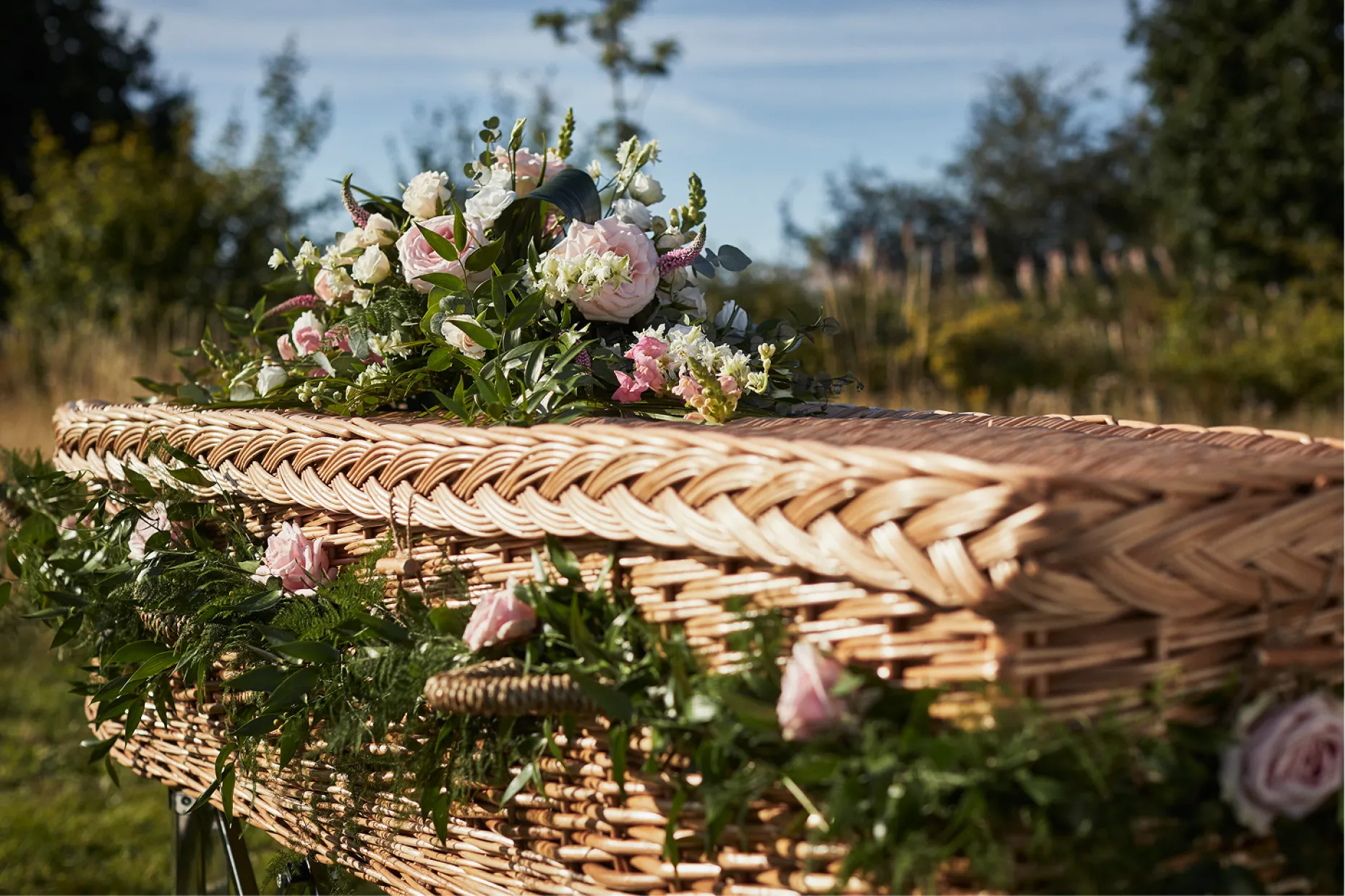 A wicker coffin outdoors with a spray of flowers on top, surrounded by garden greenery.