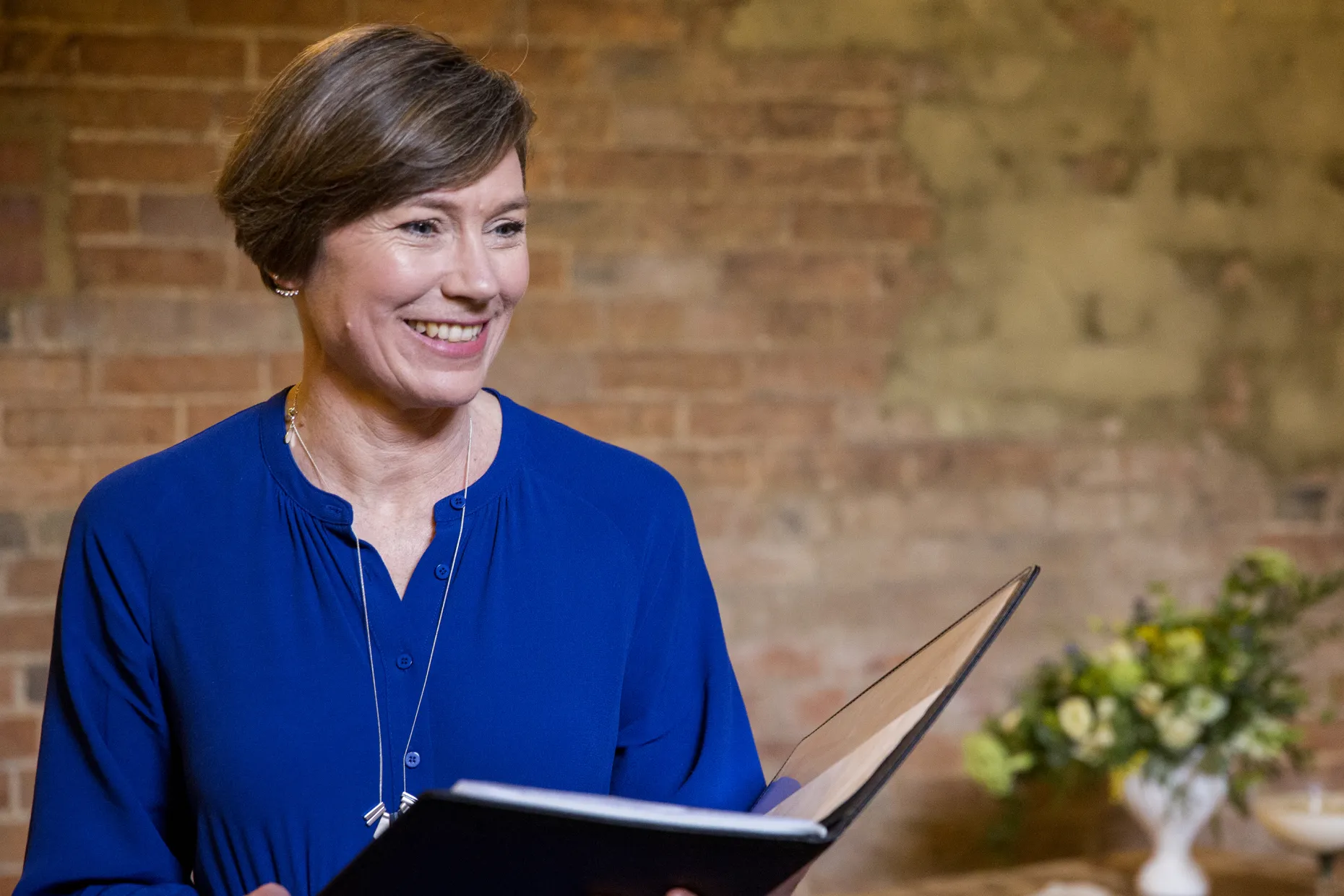 A woman in a blue dress holds an open folder and speaks, standing in front of brickwork.