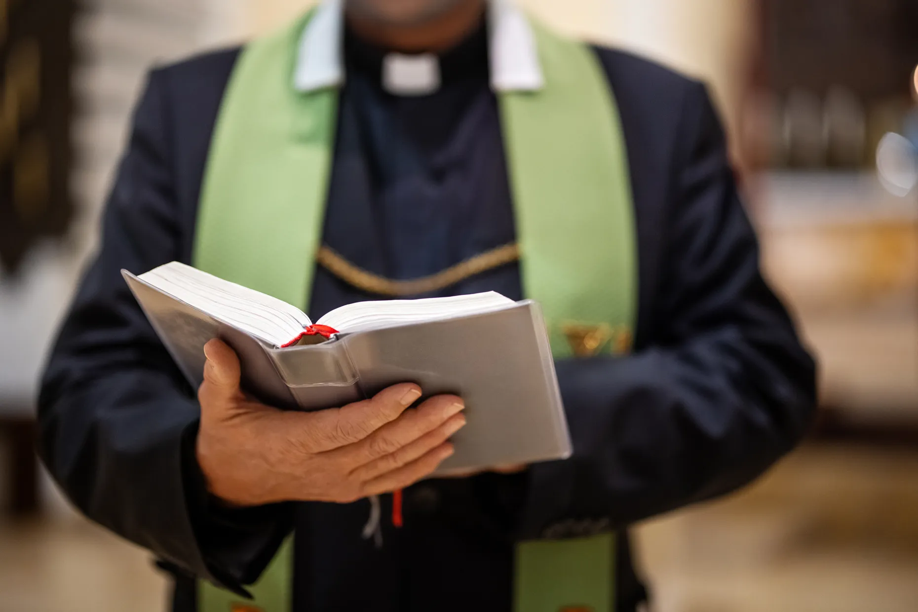 A priest in a clerical collar and green stole holds an open book in both hands.
