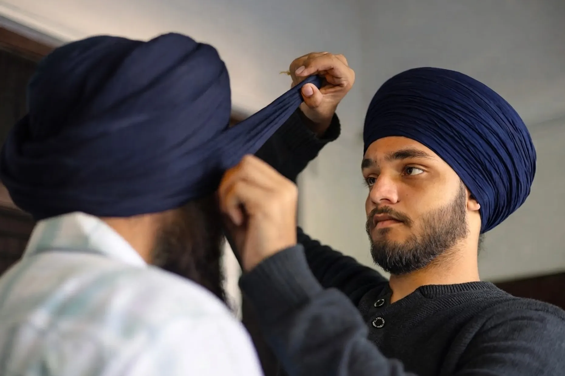 One man helps another wrap a dark blue turban, the two standing face to face indoors.
