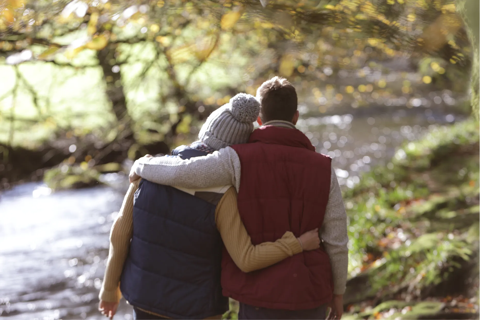 Two people in warm coats walk arm in arm along a woodland path beside a stream.