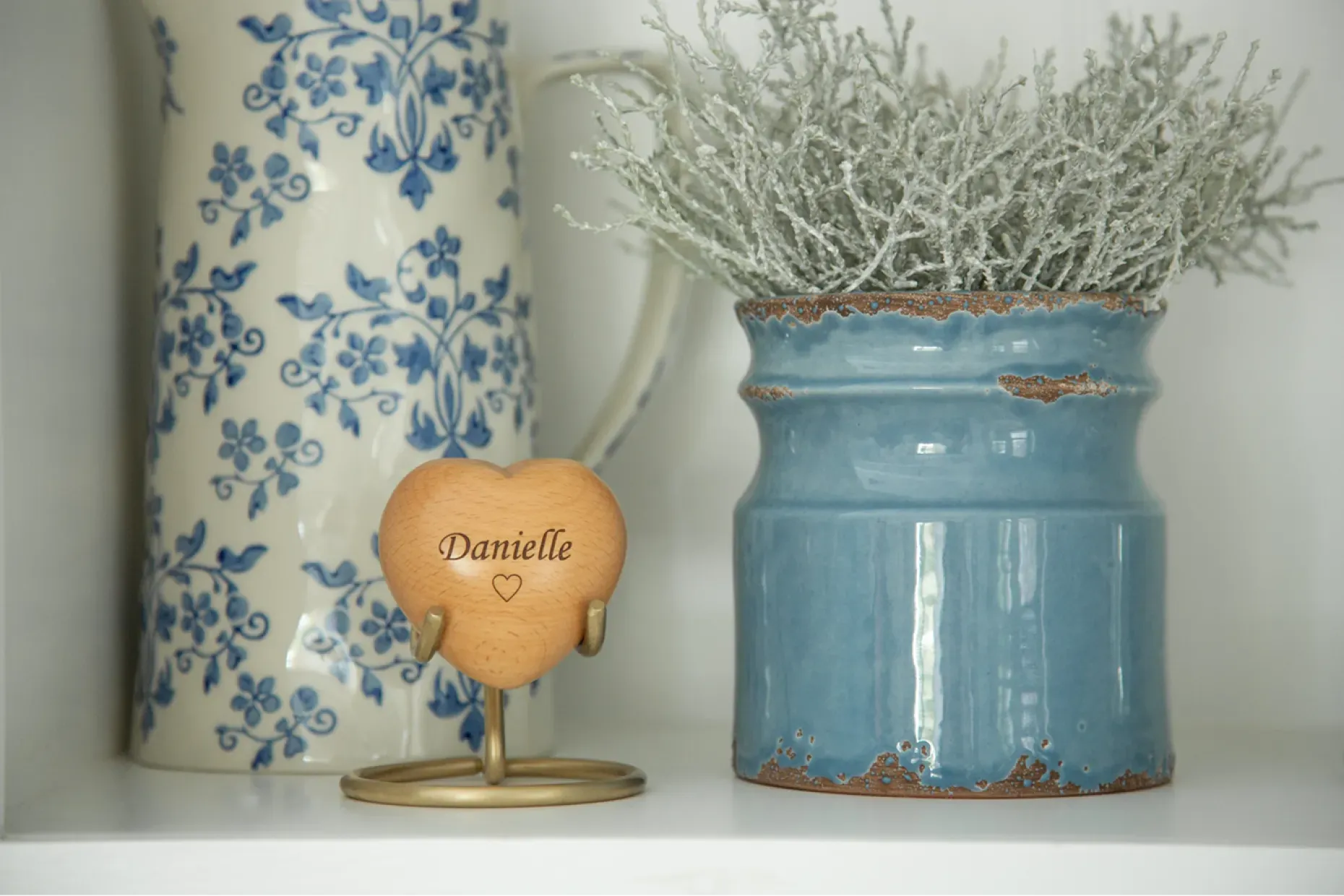 A wooden heart keepsake engraved Danielle on a brass stand, between a patterned jug and blue pot.
