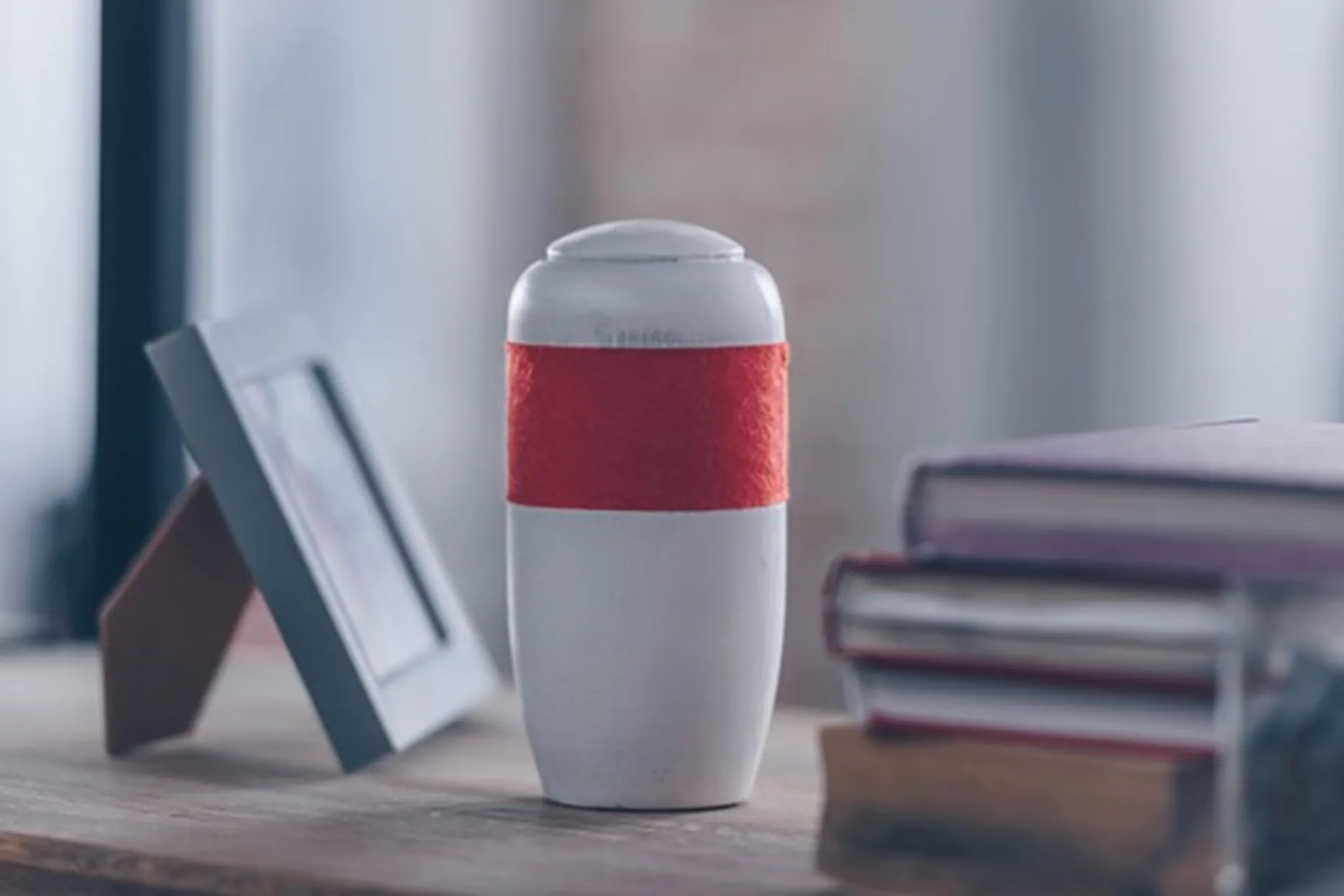 A white cremation urn with a red band, resting on a shelf beside books.